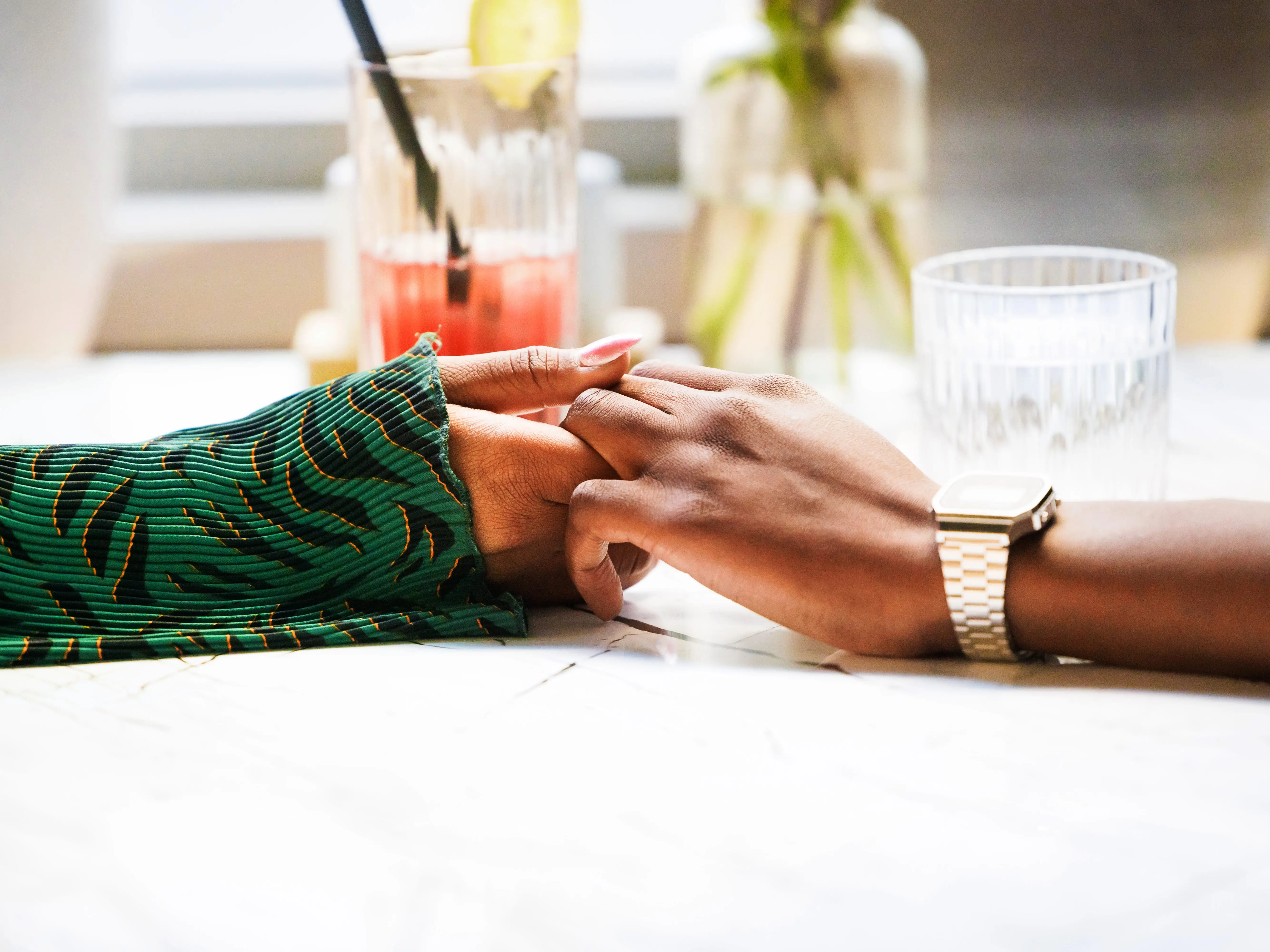 Two individuals hold hands across a table, conveying a sense of comfort and closeness in a relaxed setting.