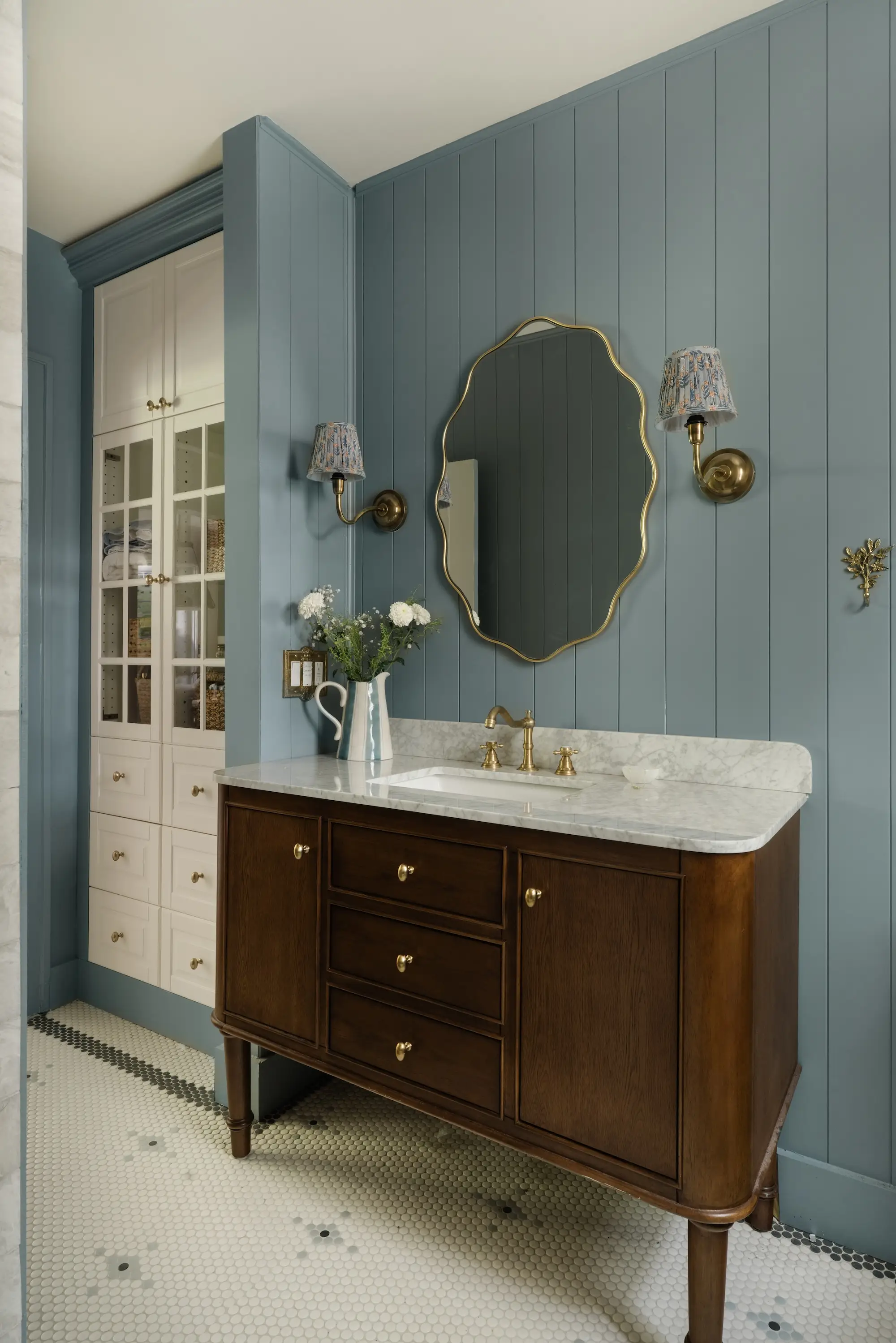 A bathroom with a wood vanity with a marble top, a curved mirror, and a white cabinet.