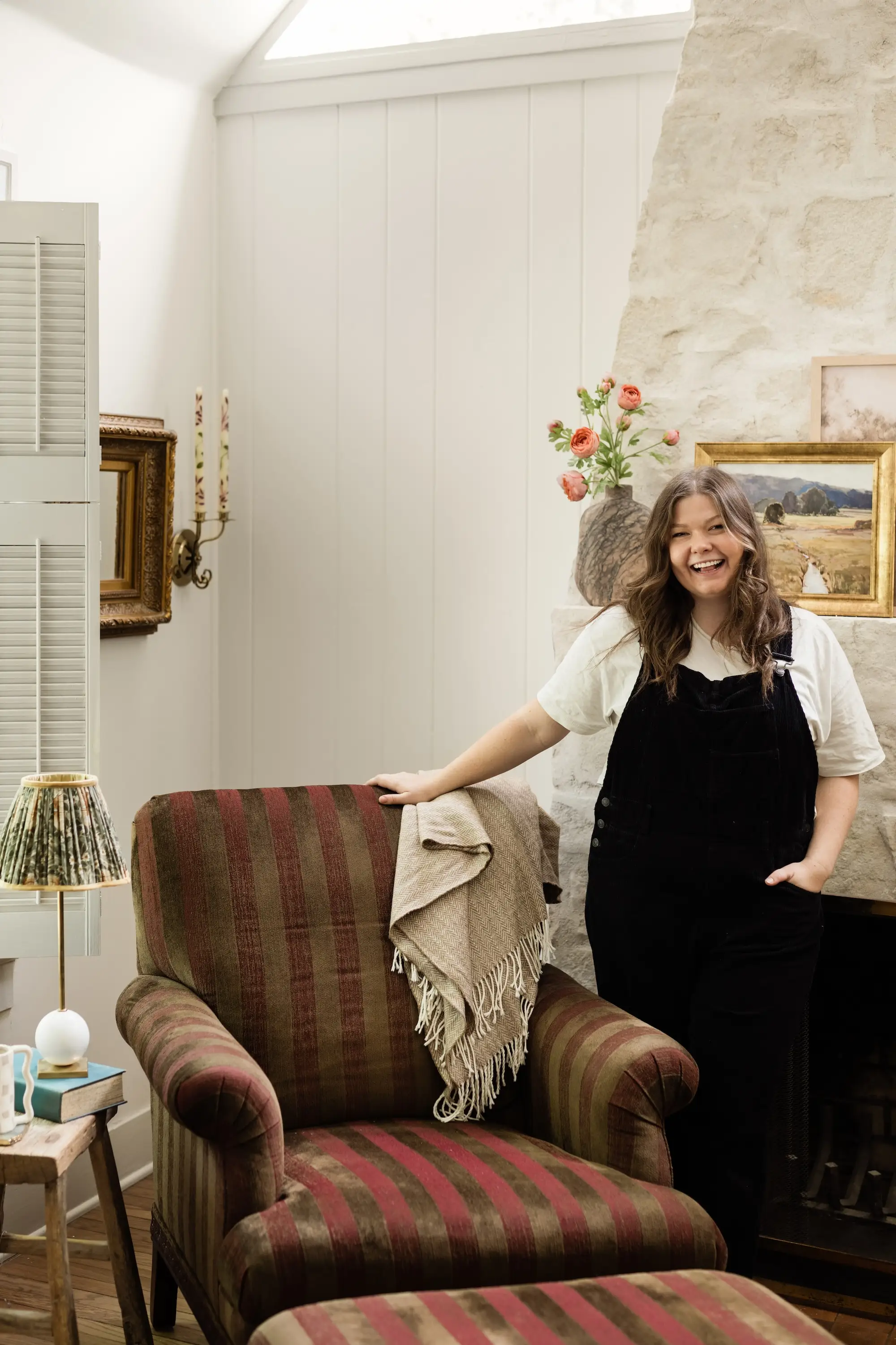 A woman smiles in front of a fireplace and rests her hand on a striped chair.