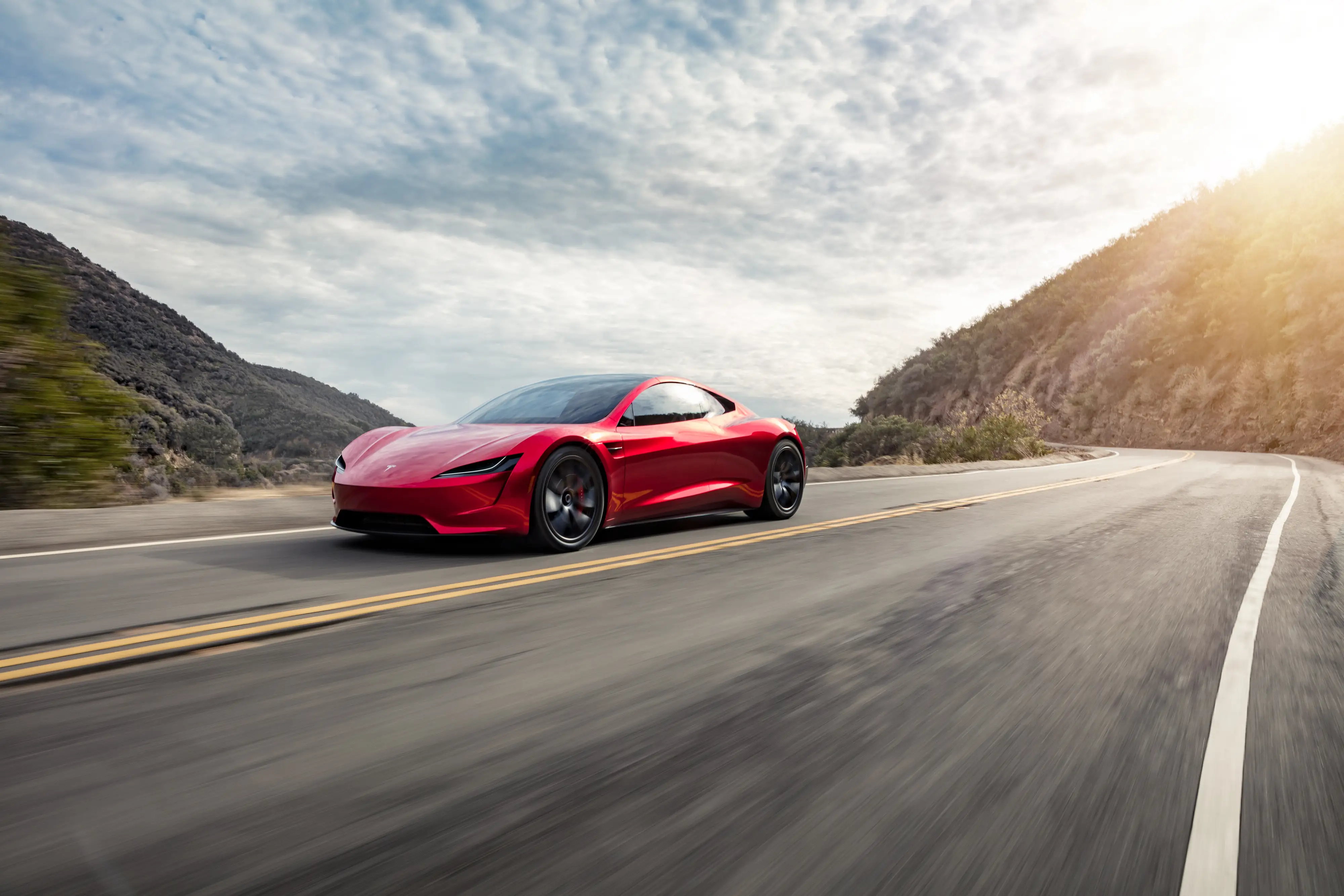 A red Tesla Roadster, a two-door sports car, drives down a hilly road.