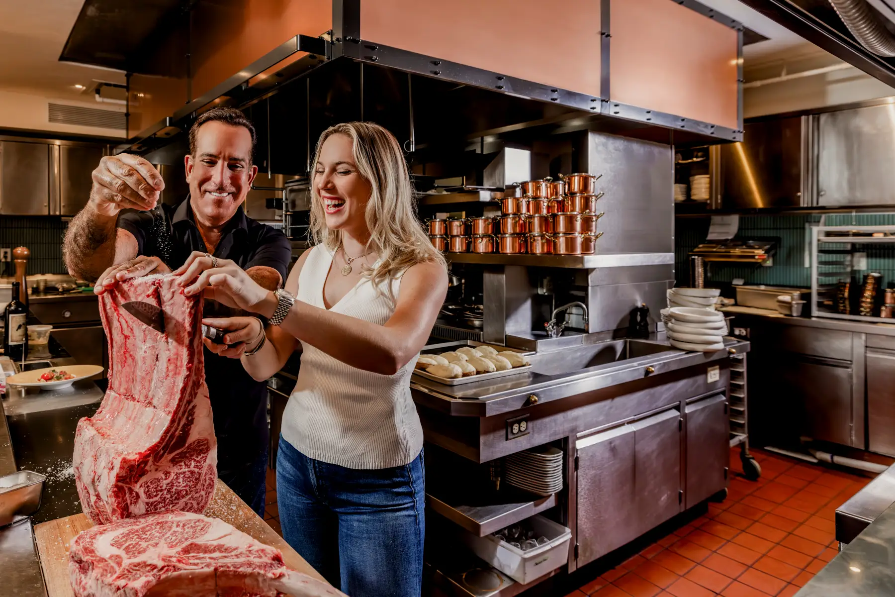 Tom and Kassidy Angelo are cooking in their restaurant kitchen, seasoning a huge slab of meat.