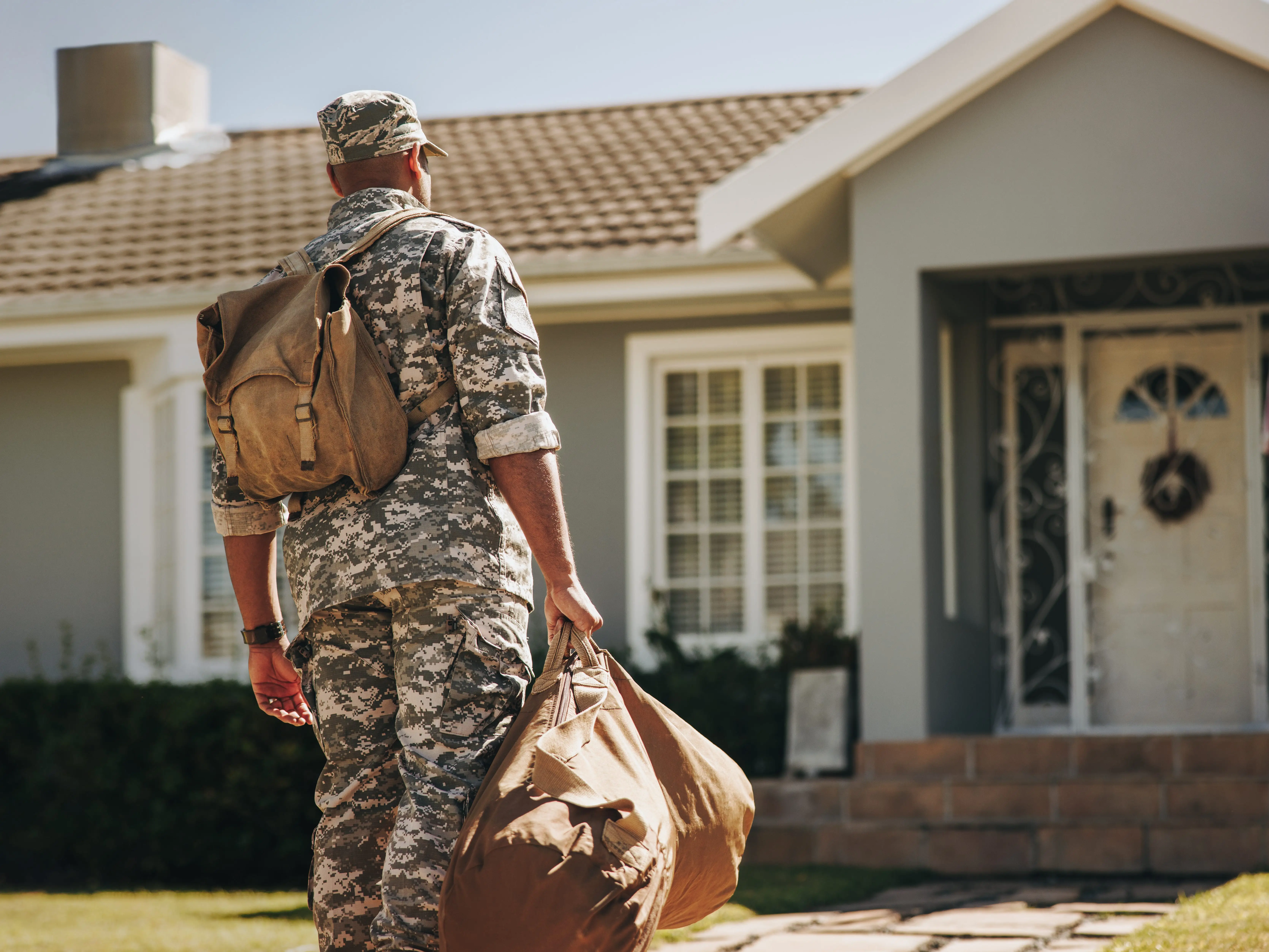 A patriotic young soldier is walking toward his house with his luggage. Rearview of an American serviceman coming back home after serving in the military.