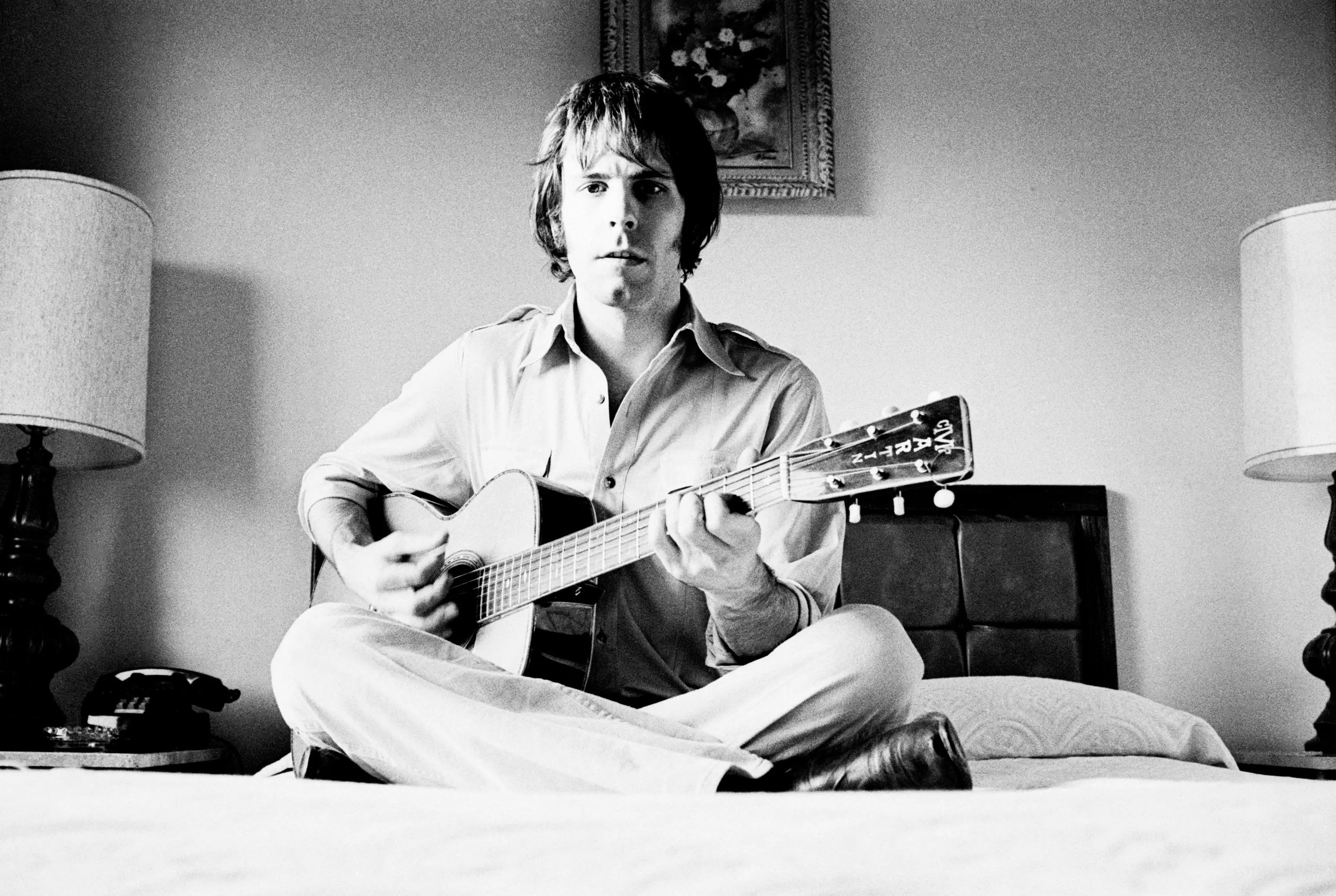Bob Weir sitting on a bed playing a guitar