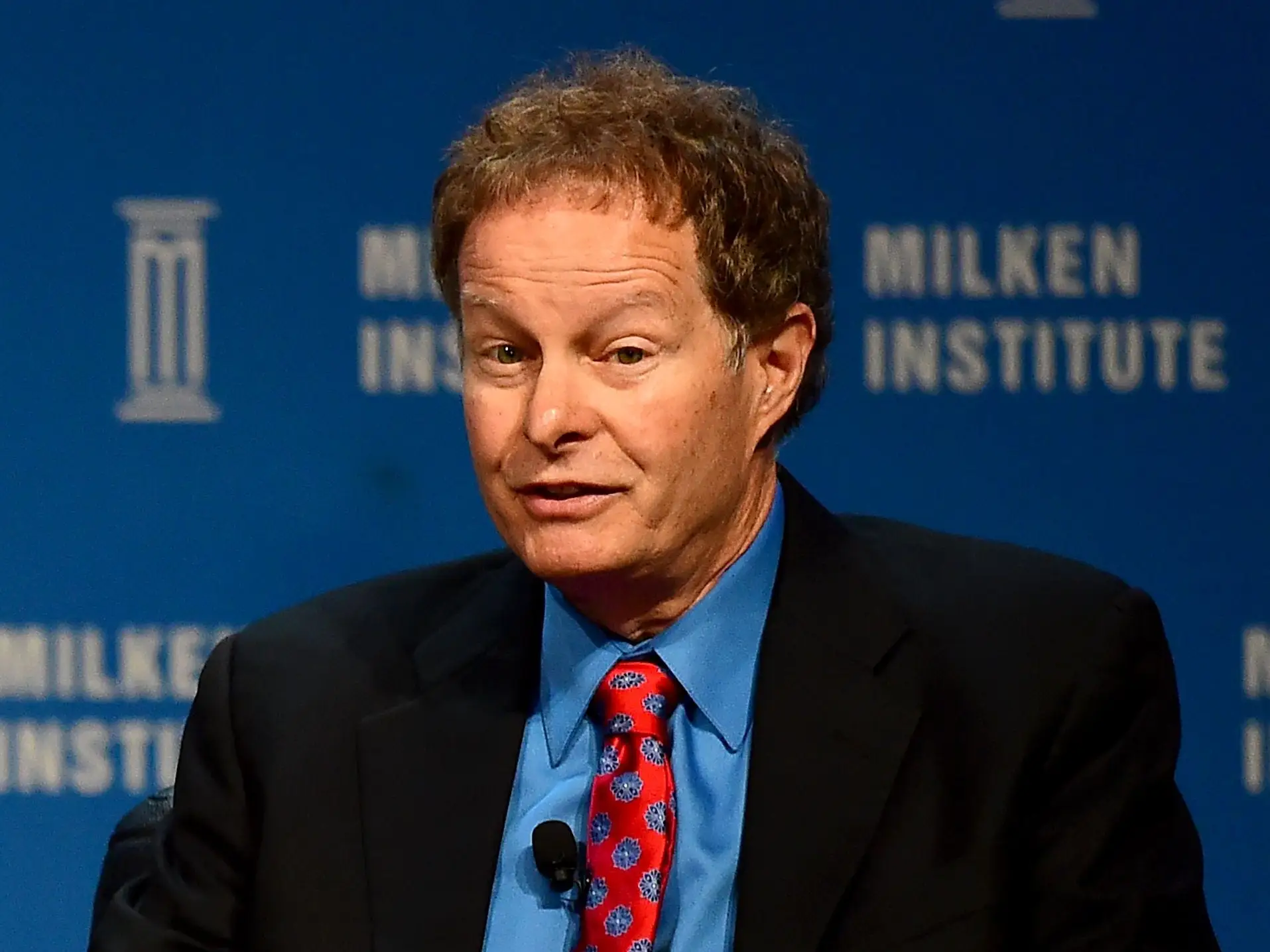John Mackey sits in a chair on stage during a presentation at the Milken Institute wearing a black suit, blue dress shirt, and red patterned tie
