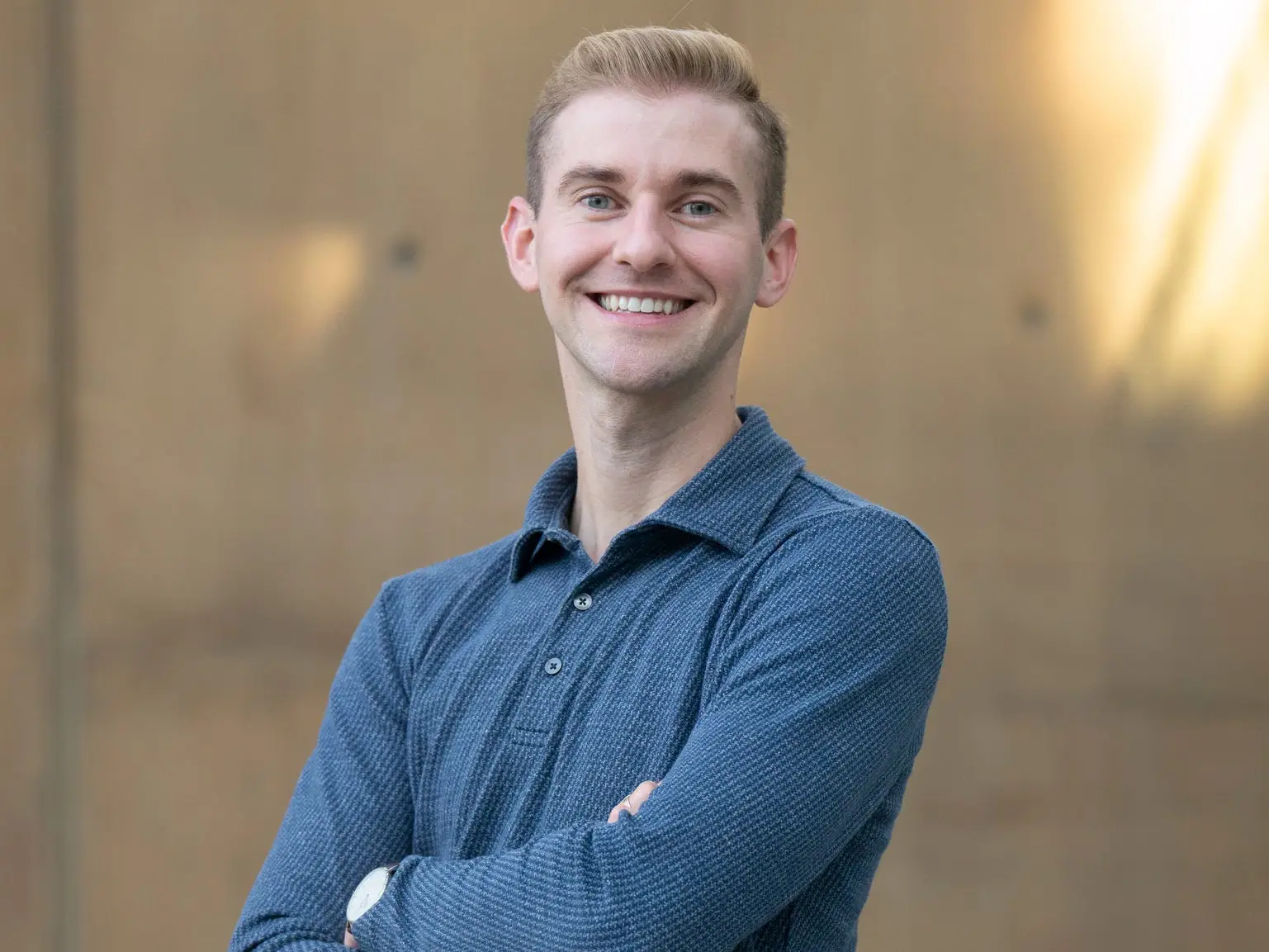 A man wearing a blue long-sleeved polo shirt smiles at the camera with his arms crossed.