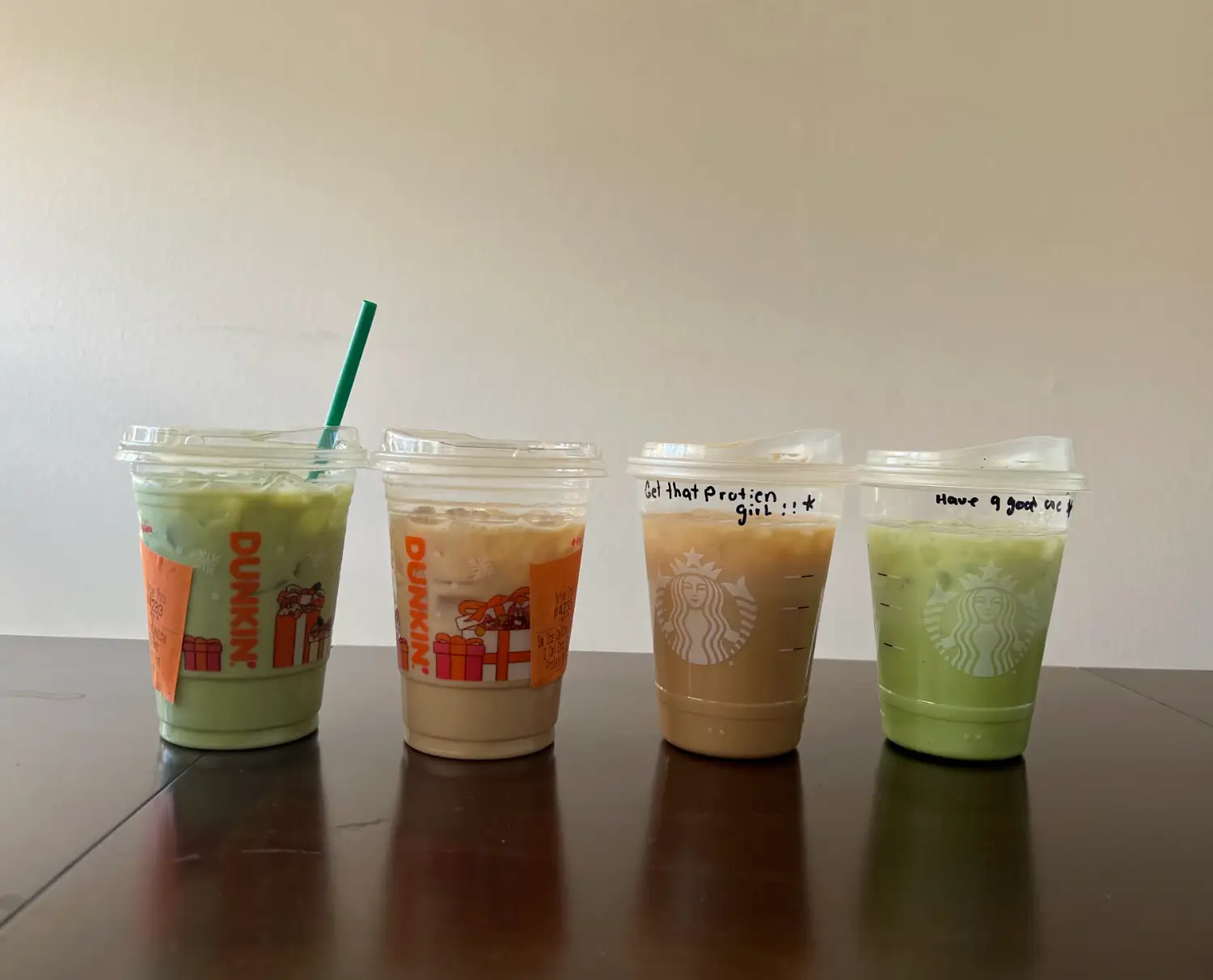 An iced protein matcha from Dunkin, a chocolate-caramel protein latte from Dunkin, a caramel protein latte from Starbucks, and an iced protein matcha from Starbucks, lined up on the writer's table.