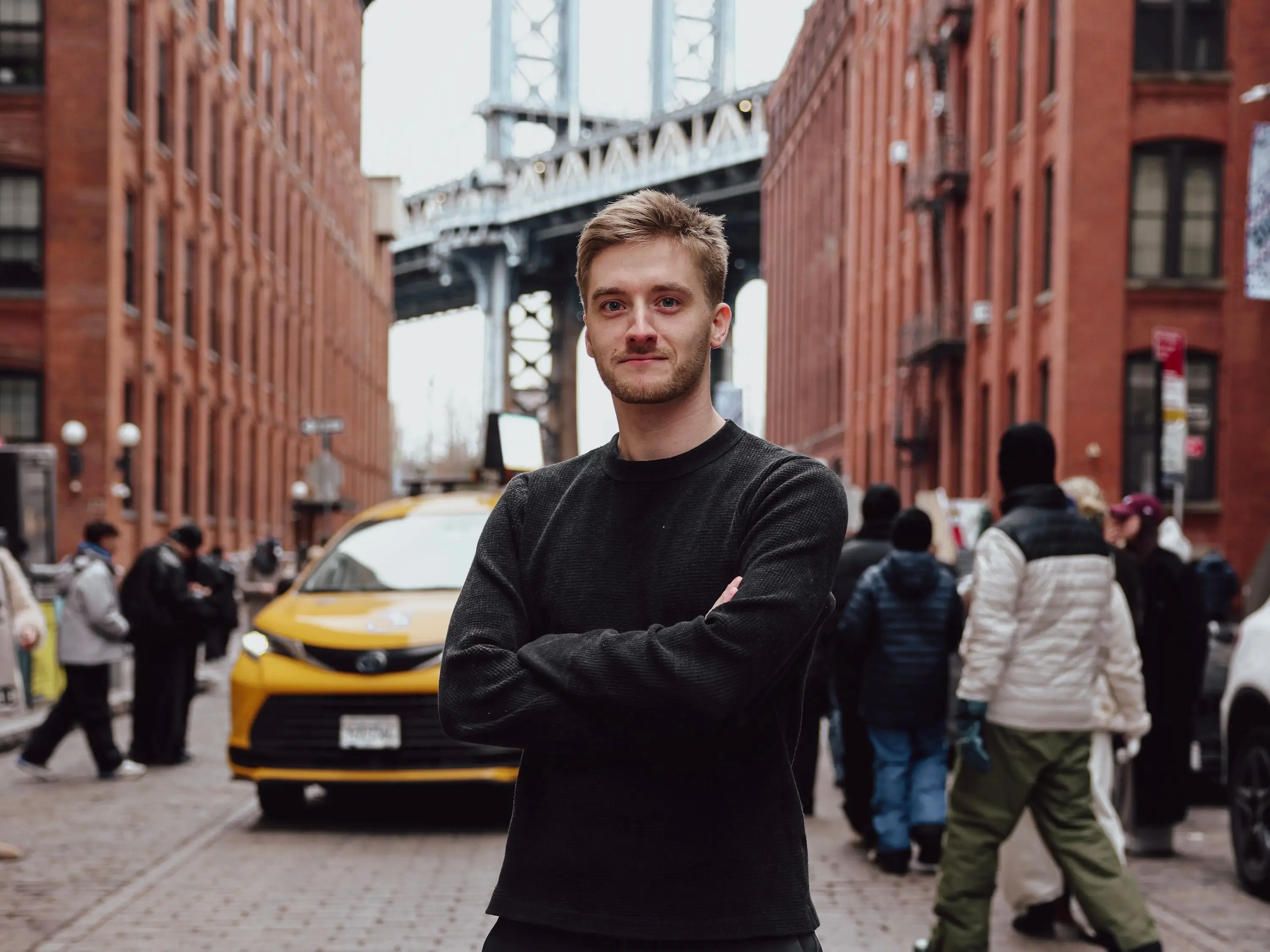 A man stands with his arms crossed on a cobblestone street in Brooklyn's Dumbo neighborhood, with the Manhattan Bridge rising between red-brick buildings behind him.