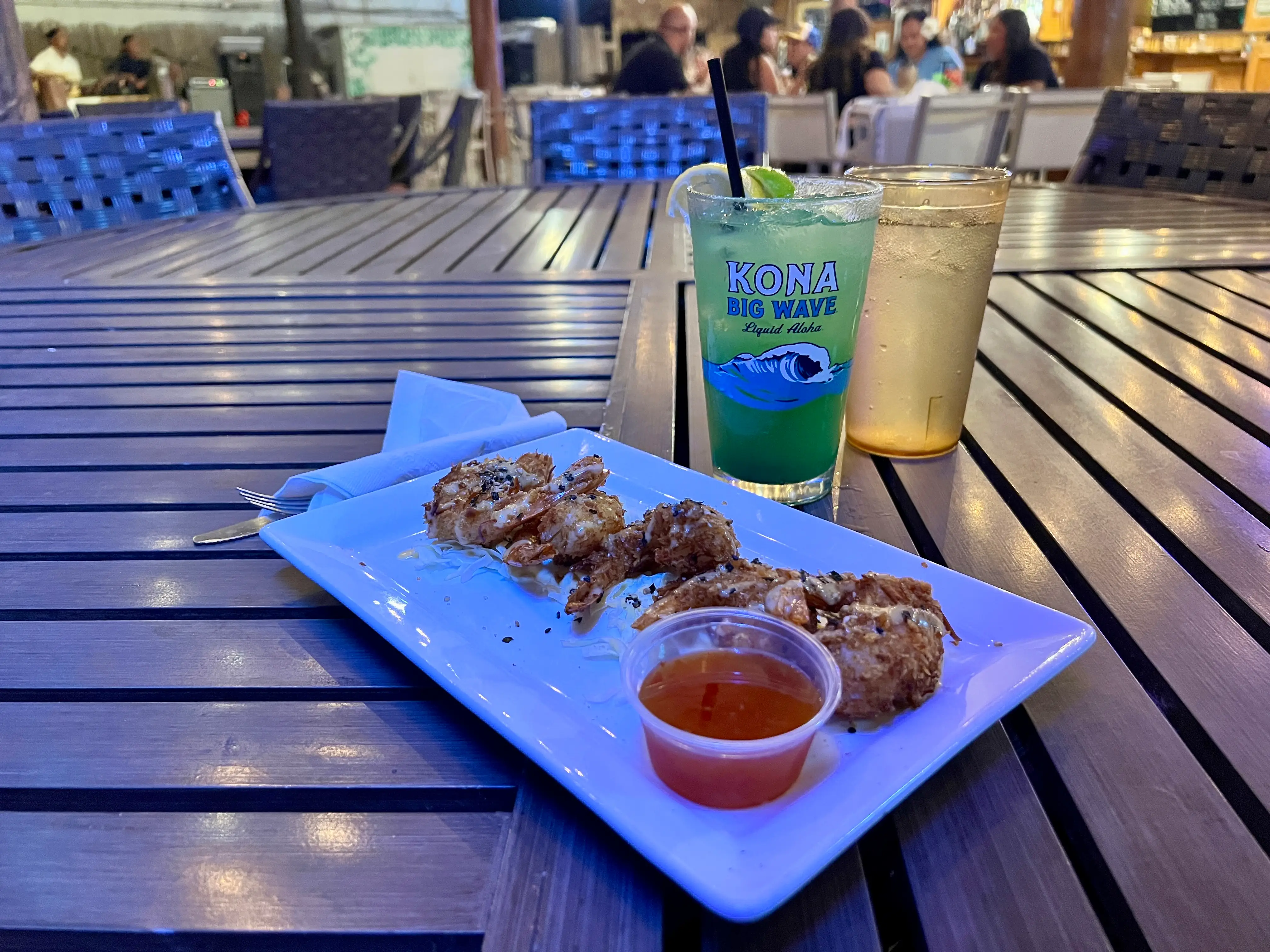Plate with chicken pieces, sauce next to Kona Big Wave glass on table in evening