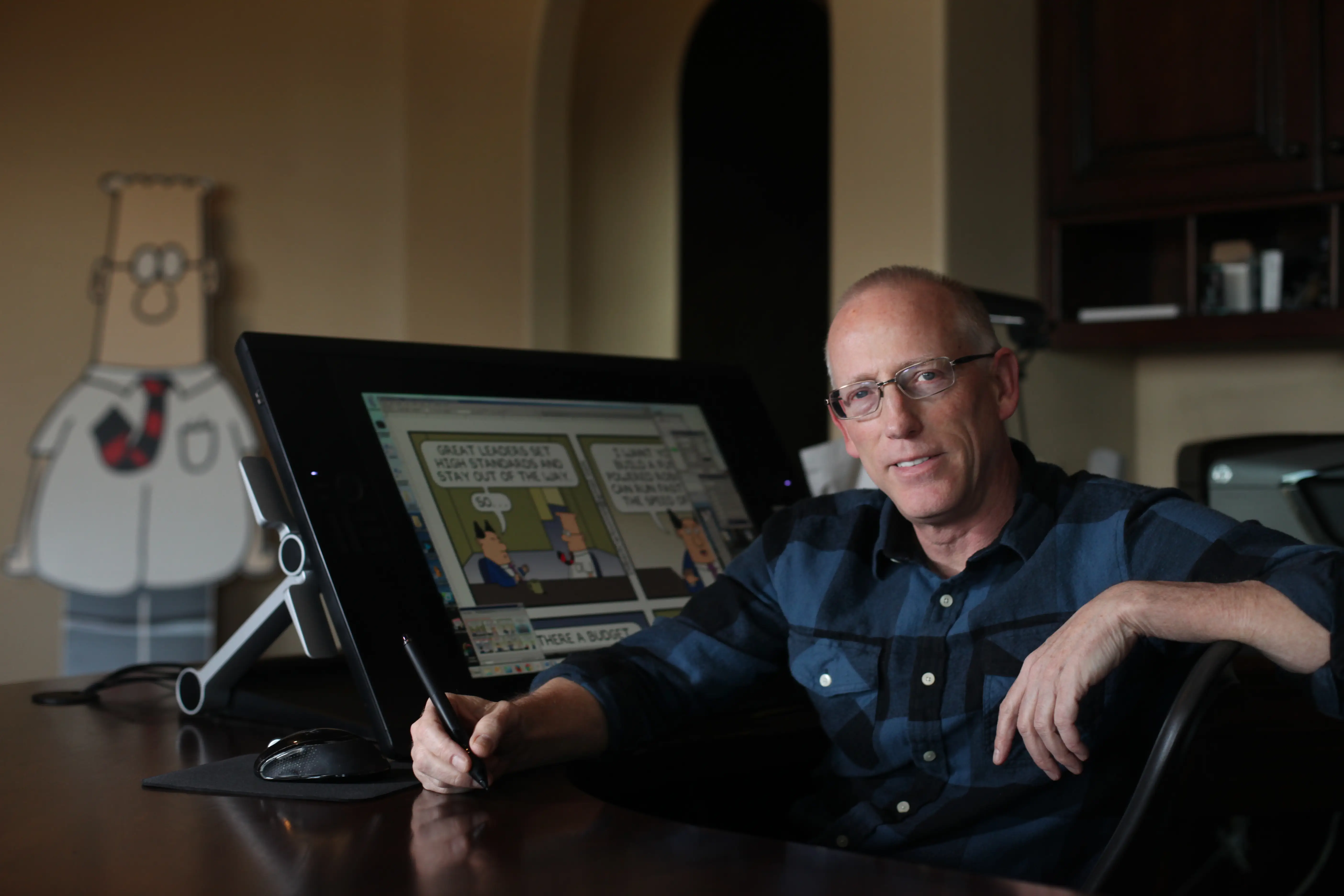 Scott Adams at a desk sketching Dilbert with a Dilbert cutout behind him