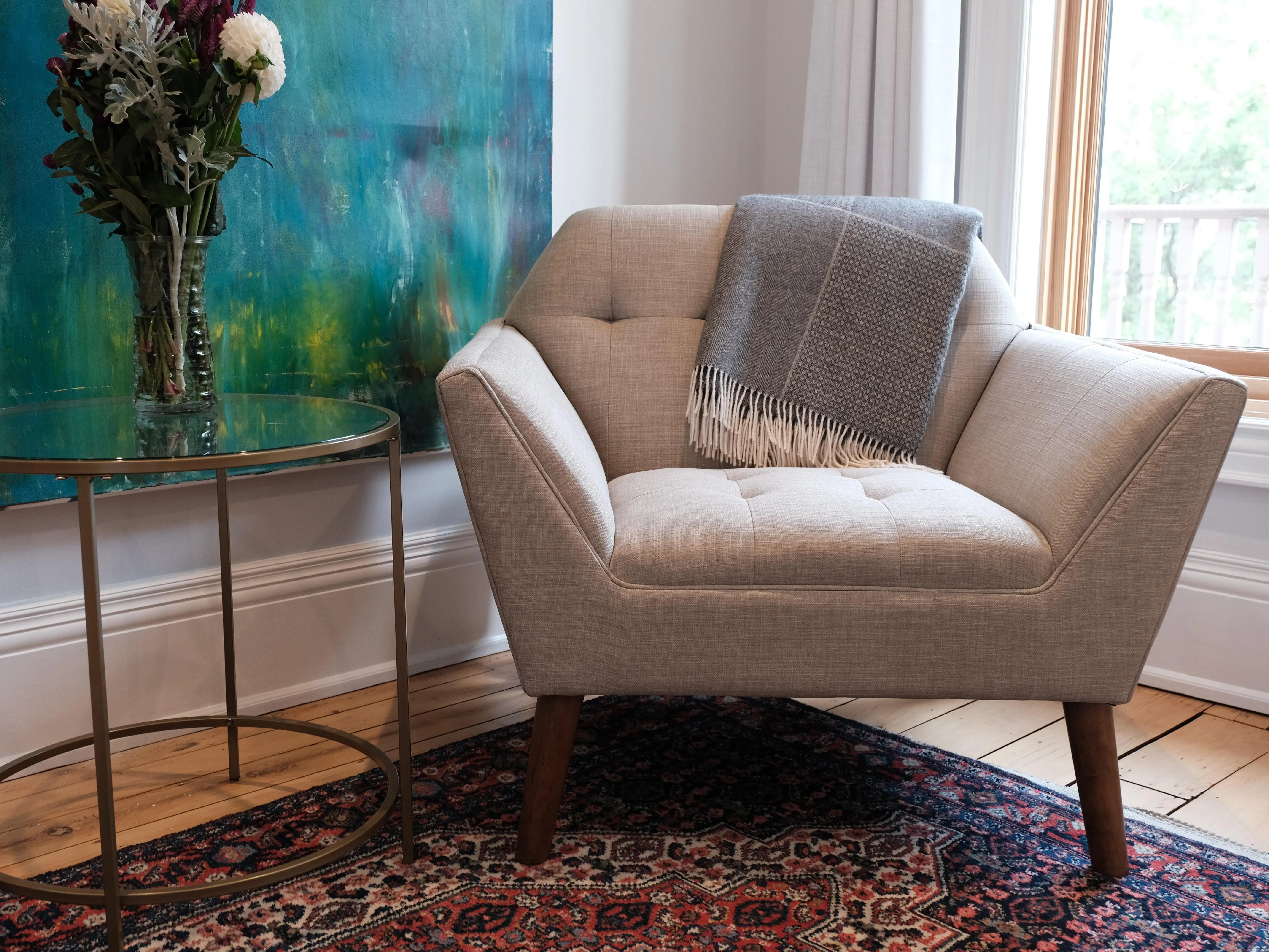 An accent chair and table adorned with a blanket and flowers, respectively. There's a textured, patterned rug on the floor and blue-green artwork on the wall.