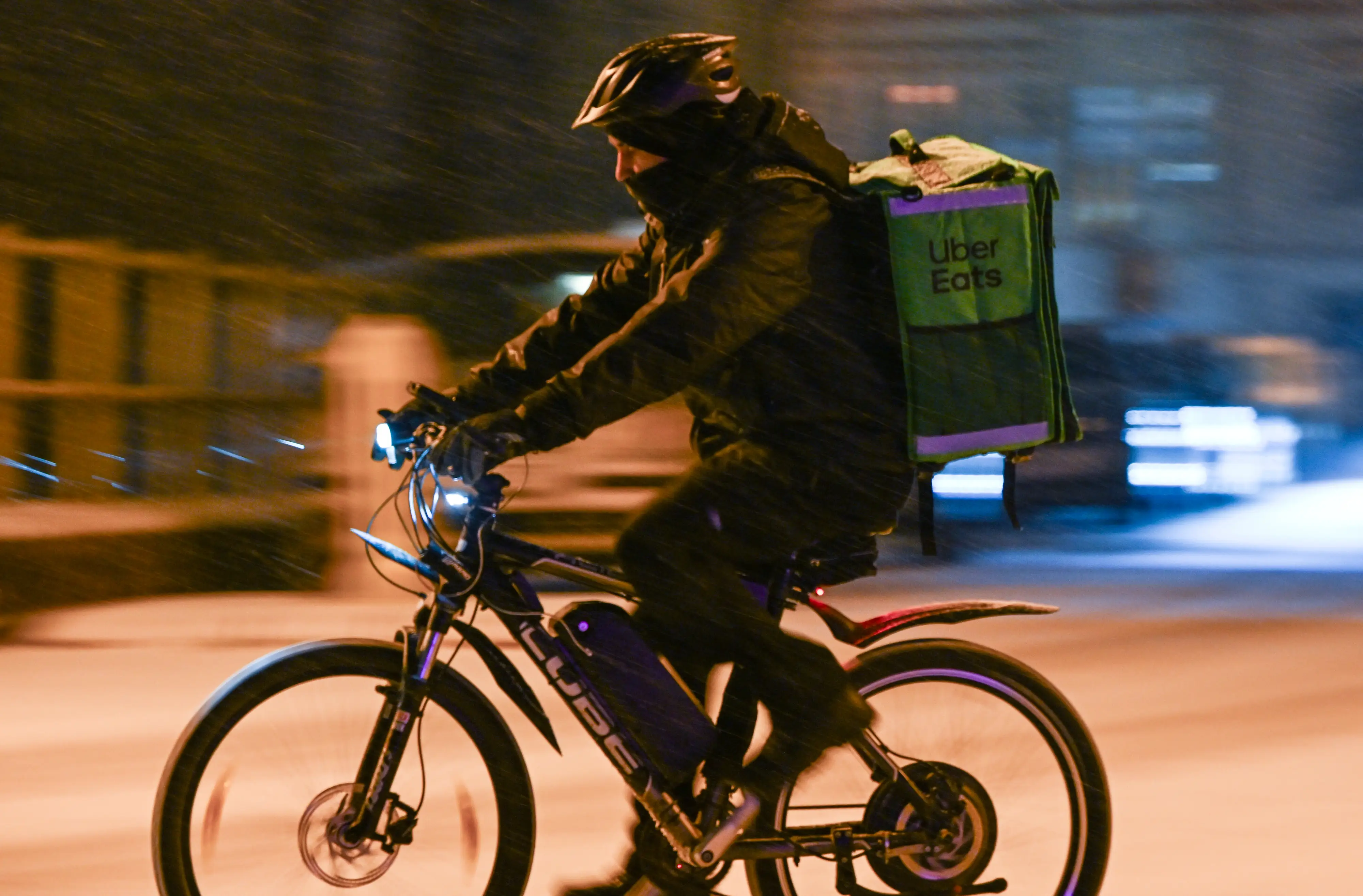 An Uber Eats delivery worker rides an e-bike while wearing a green Uber Eats backpack and a helmet at night in the snow.