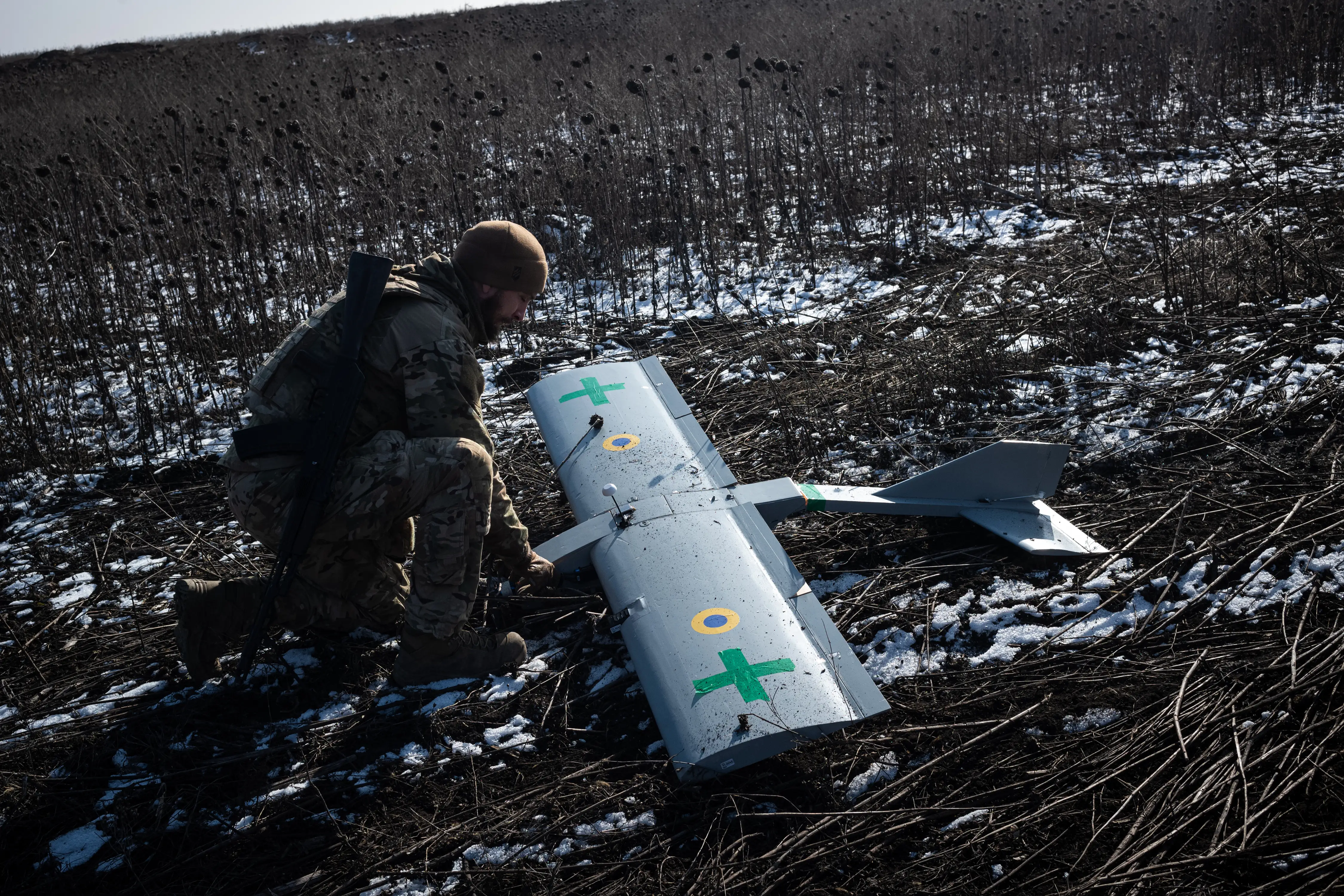 A man kneels beside a large grey drone in muddy and snowy ground
