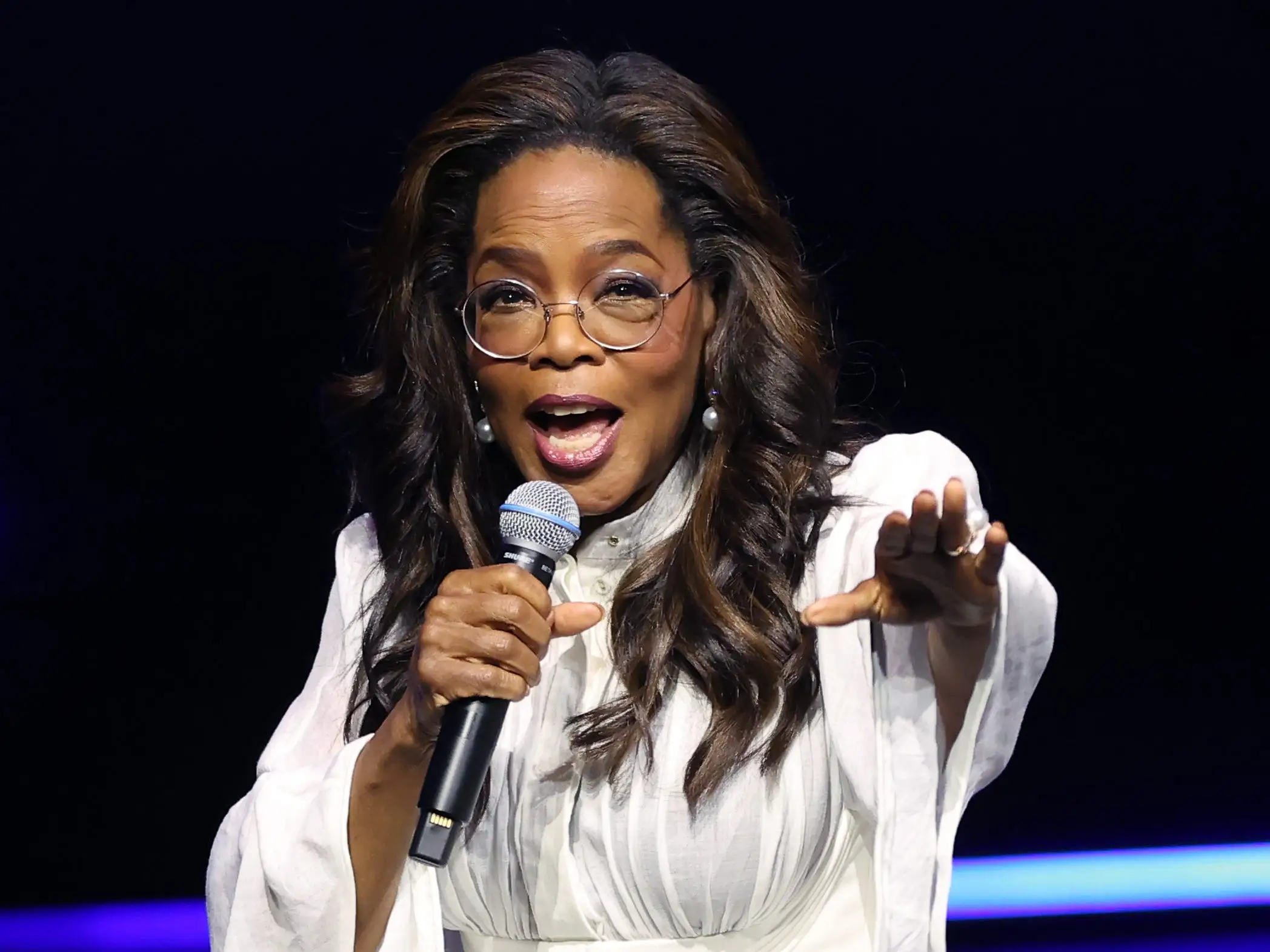 Oprah Winfrey on stage in a white dress with a microphone, holding her hand out to the audience.