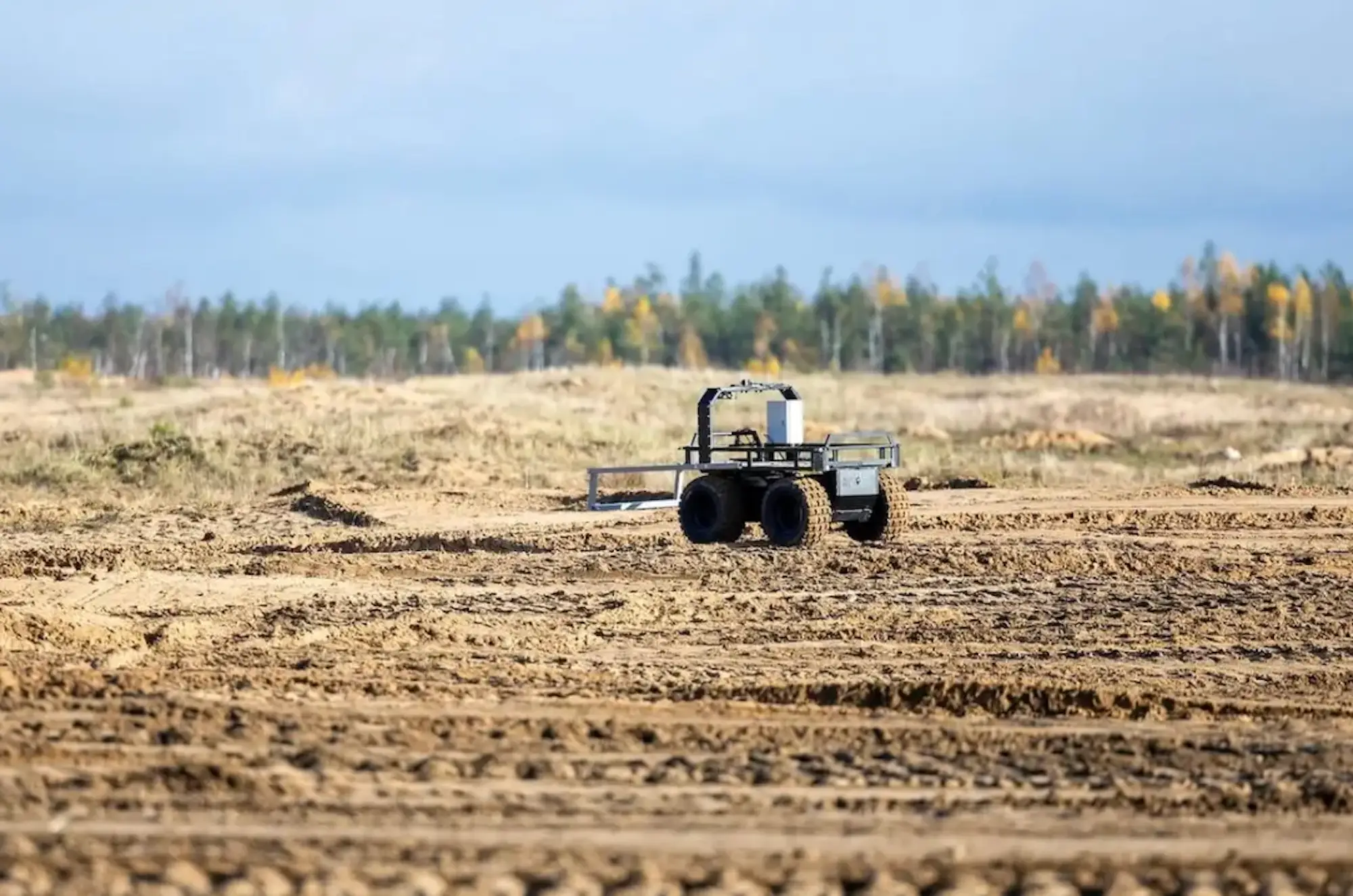A four-wheeled robot on a golden muddy ground with trees and a cloudy sky behind