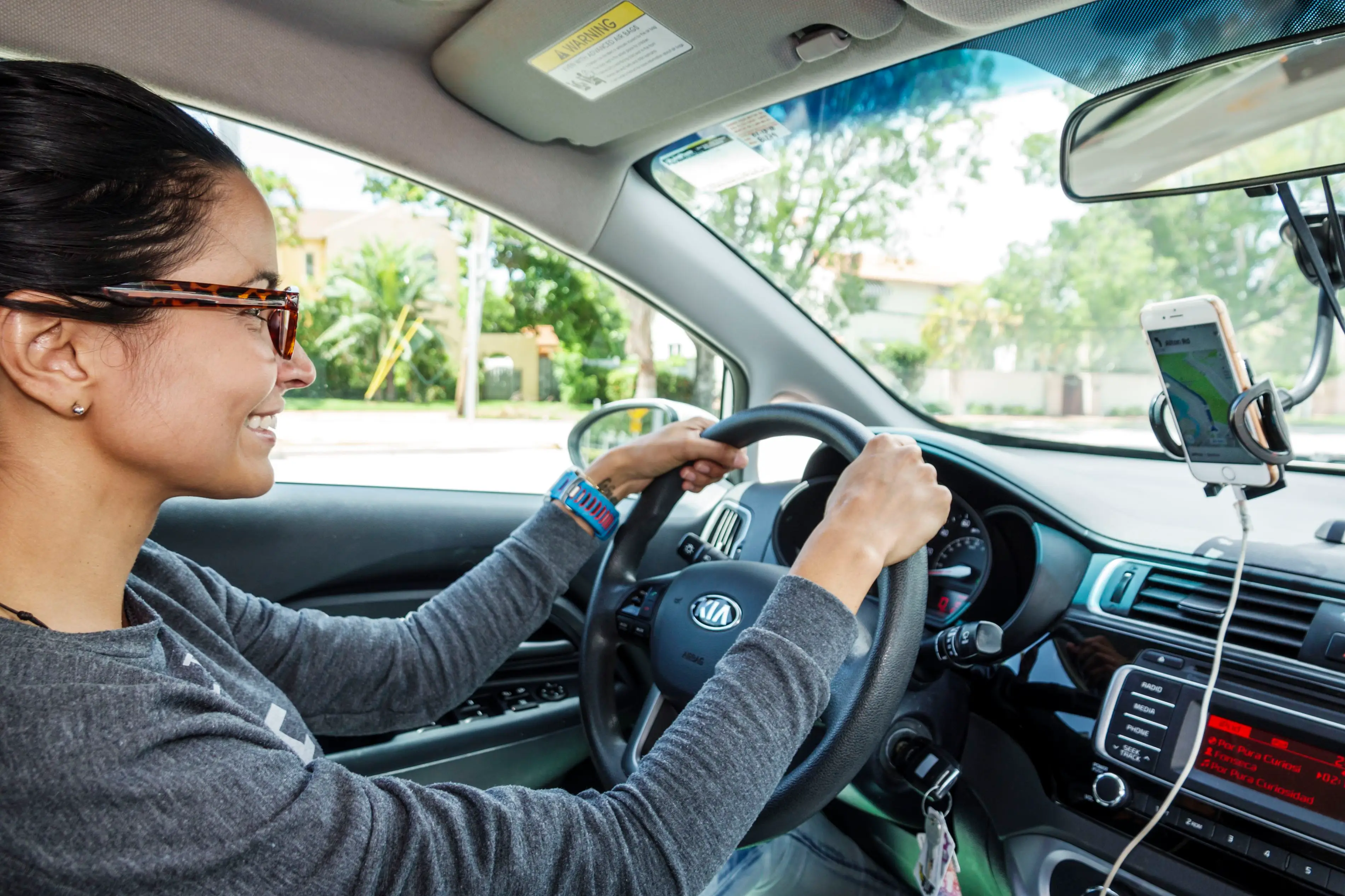 A woman wearing glasses holds the steering wheel of a Kia car while smiling. To her right, a smartphone sits in a holder attached to the inside of the windshield and plugged into a charger.