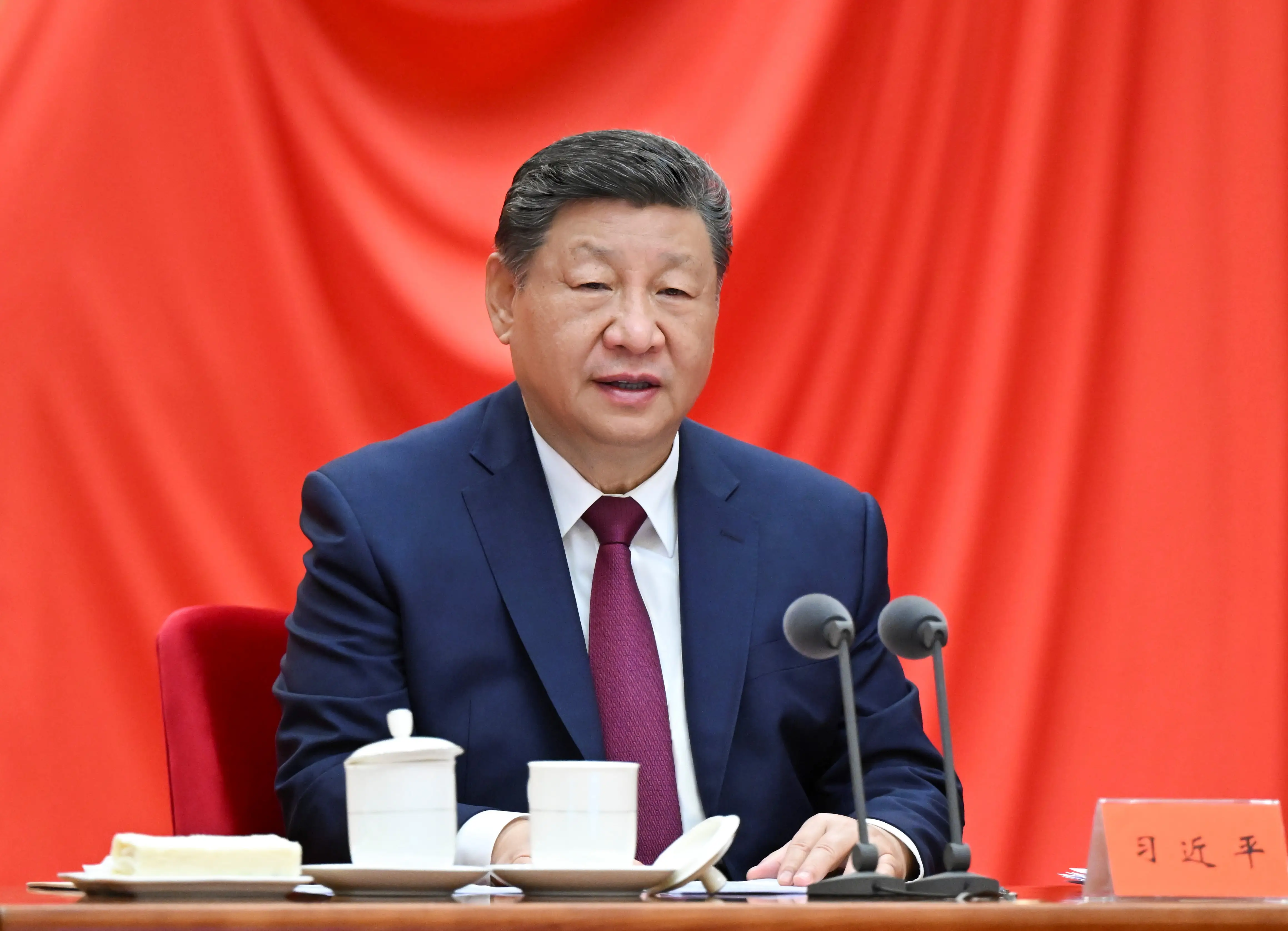 Xi Jinping sits at a microphone in front of a red backdrop