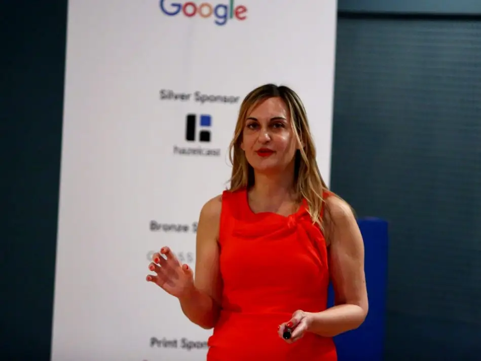 A woman in a red dress gestures while speaking at a conference, standing in front of a sponsor banner displaying the Google logo.