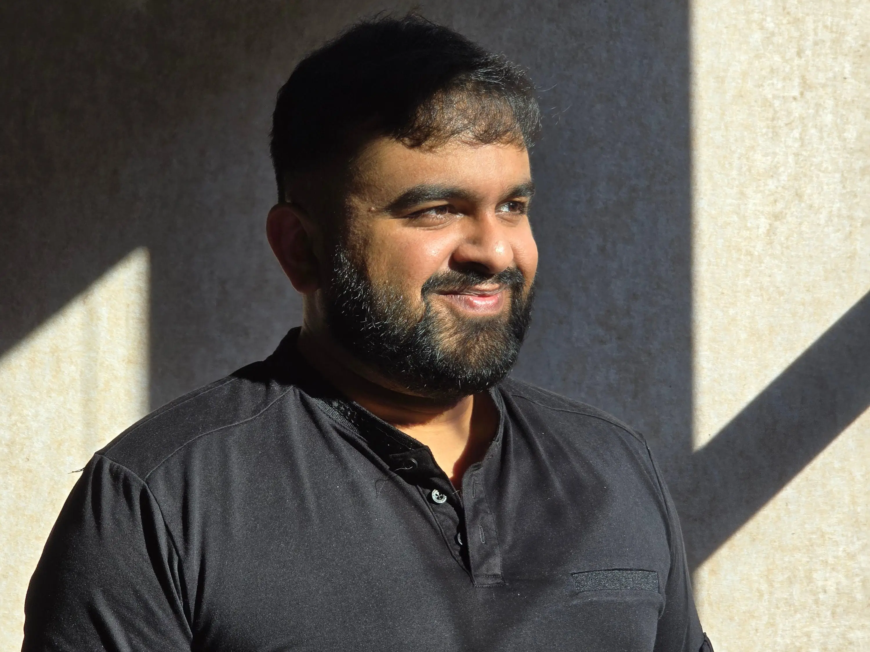 Pardeshi is wearing a black T-shirt and standing in front of a plain wall.