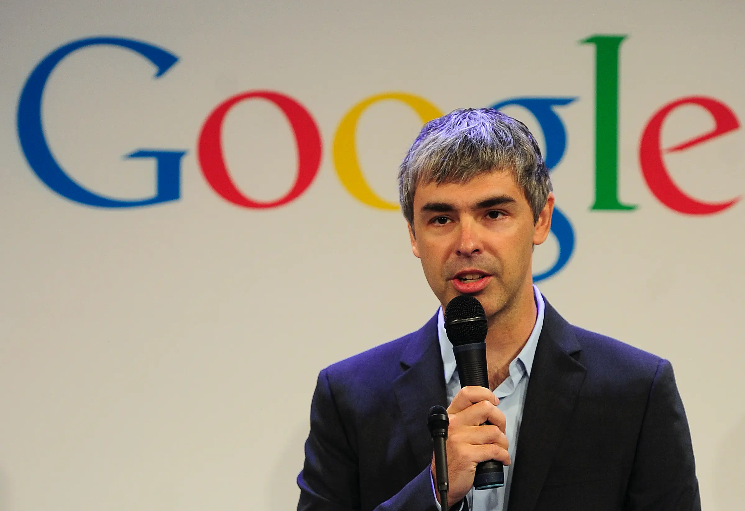 Larry Page holding a microphone standing in front of a Google backdrop.