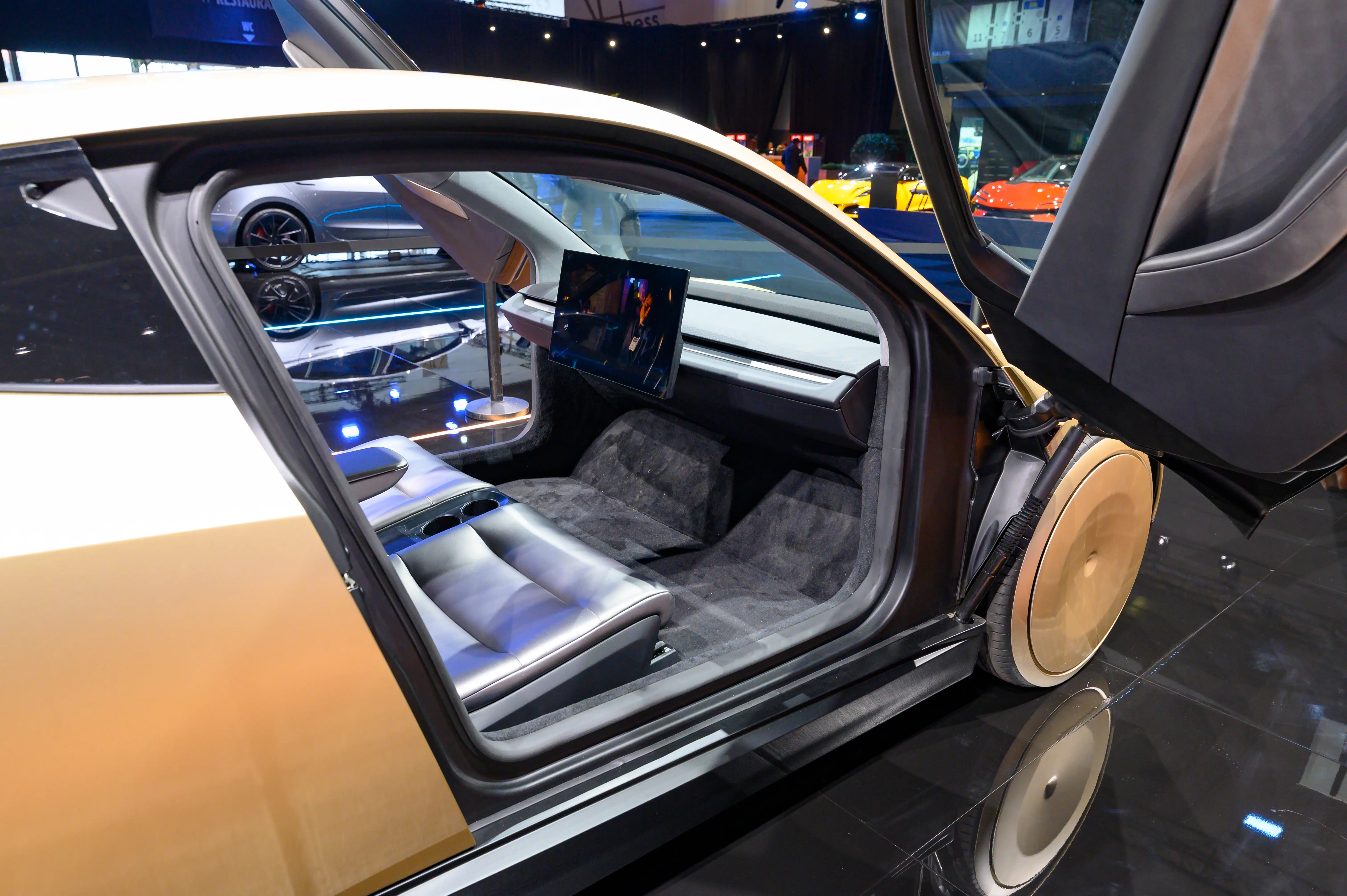 A gold Tesla Cybercab displayed at an auto show - the car has two seats, no steering wheel or pedals, a large center screen, and doors that swoop upward when opened.