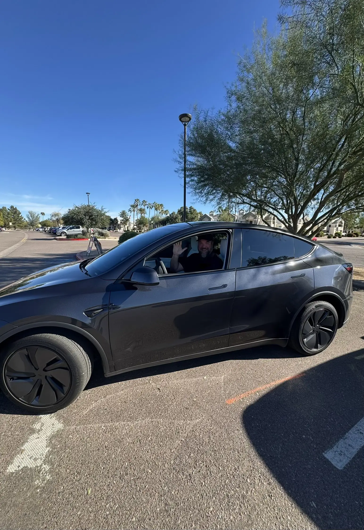 Scott Anderson, a Tesla owner, waves at the camera from the drivers seat of his brand new gray Model Y.