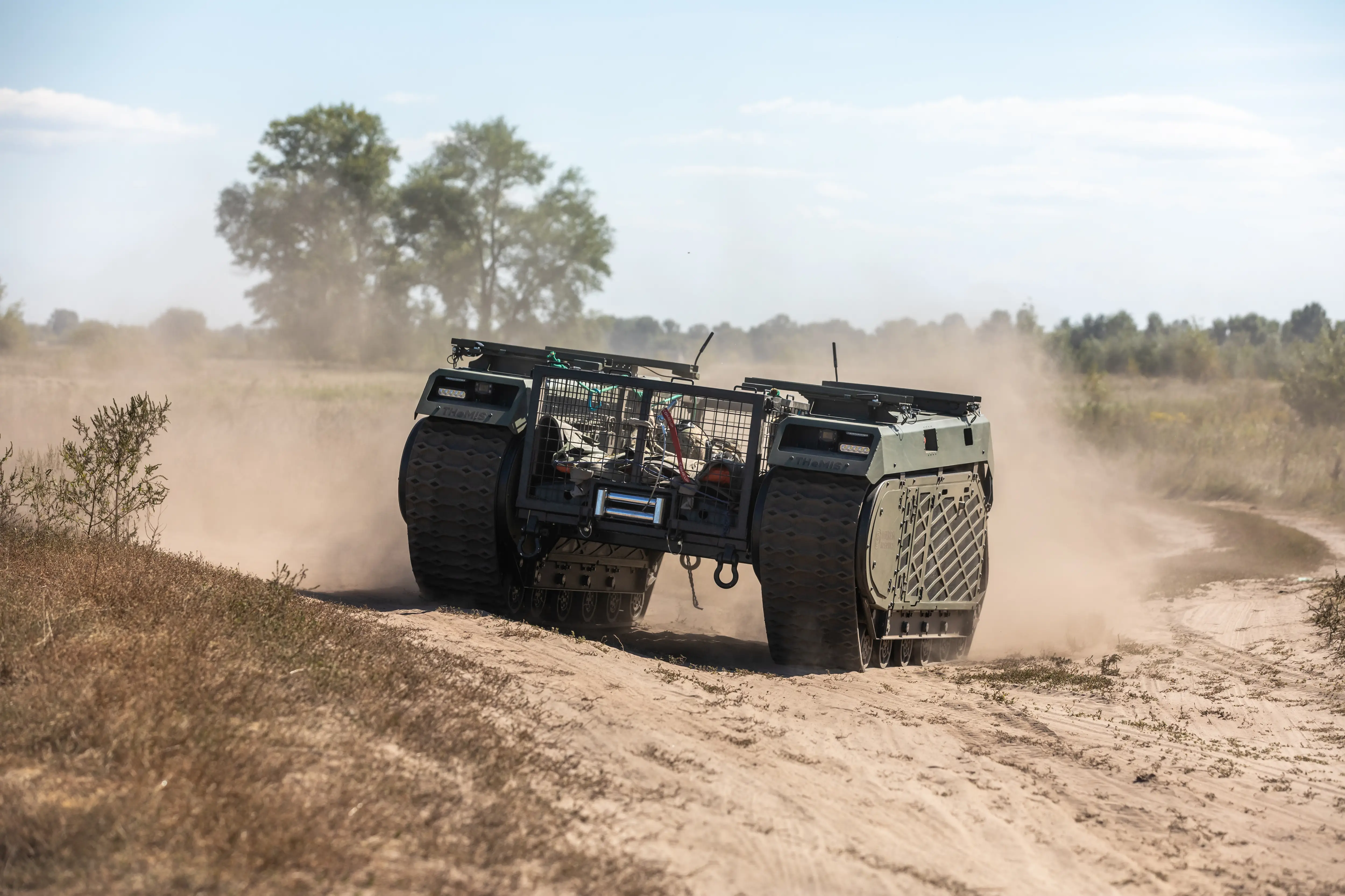 A large green robot on wheels driving on a dusty track under a blue sky