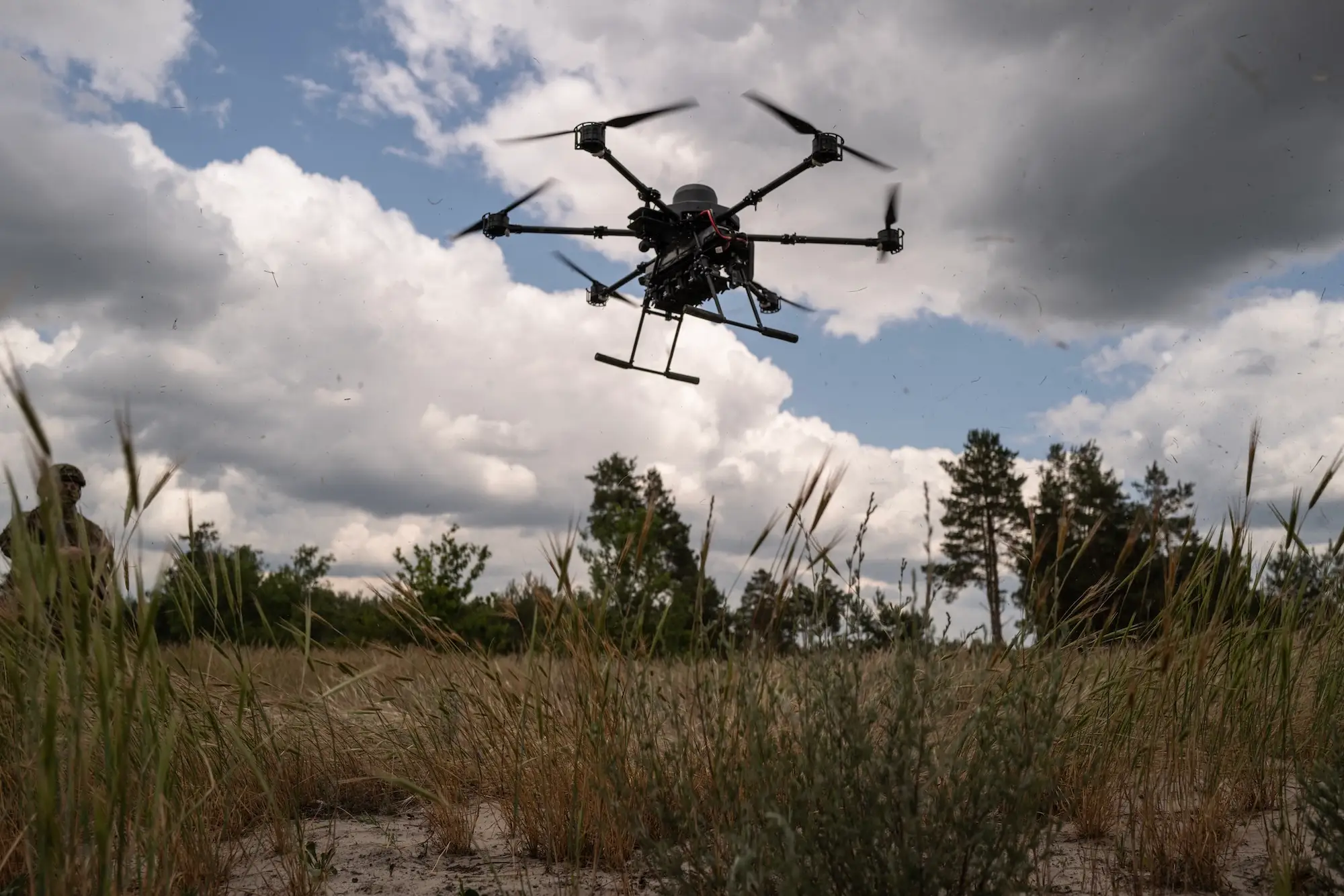 A black drone with mutiple rotors in the air above a grassy ground with a blue but cloudy sky