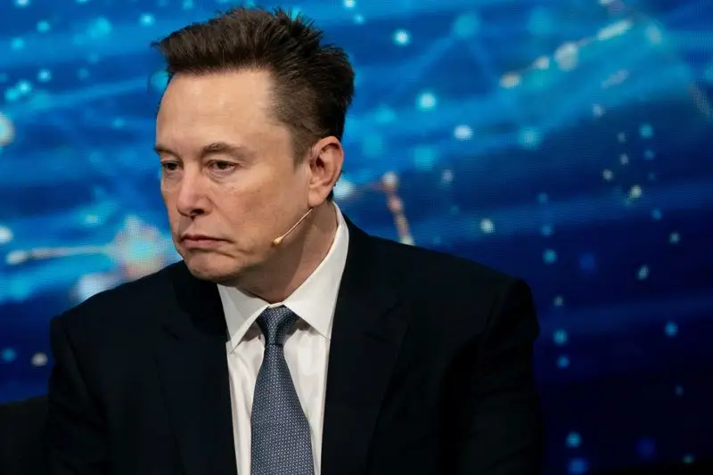 Elon Musk, dressed in a black suit and a blue tie, looks to his right during an on-stage interview.
