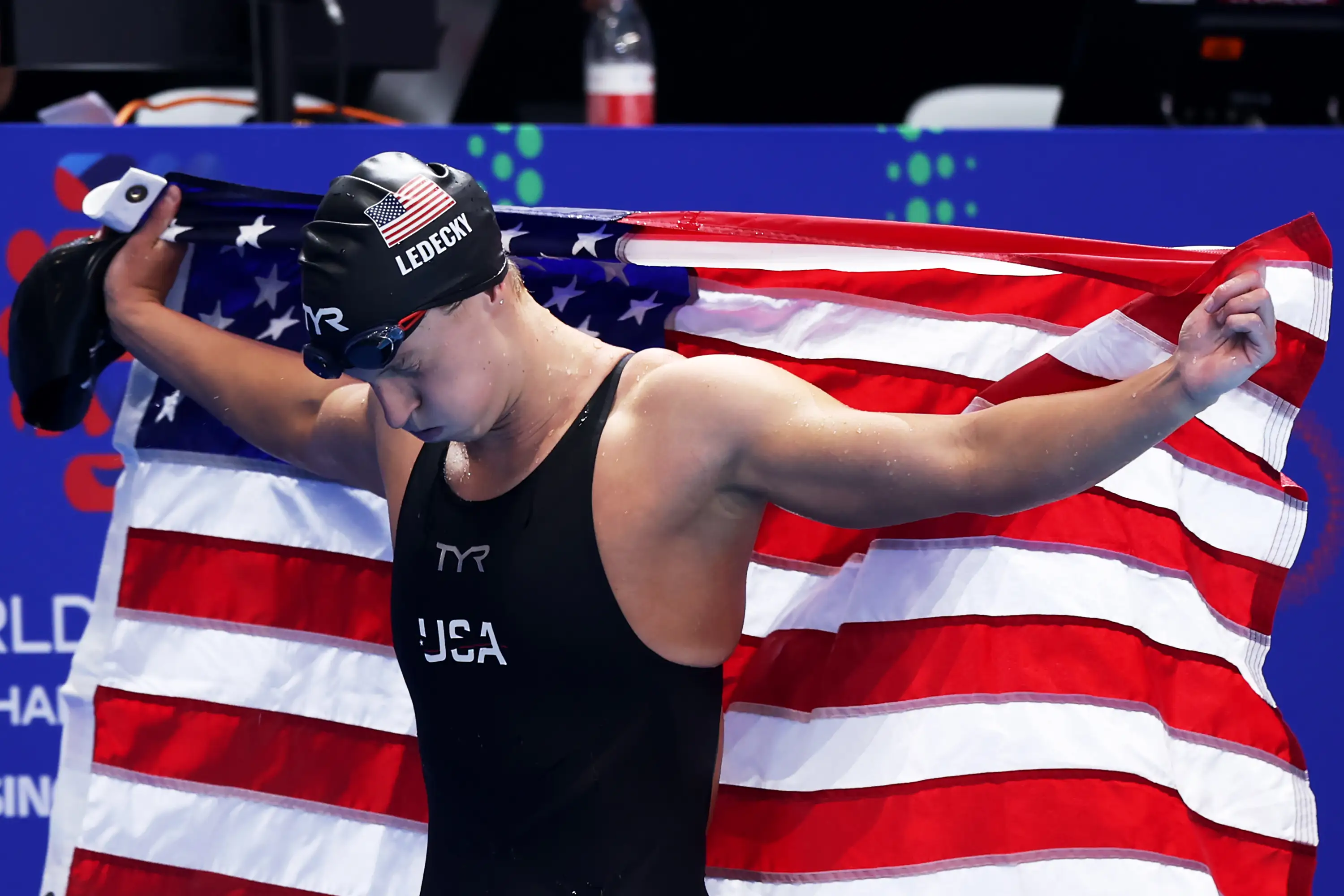 Olympic swimmer Katie Ledecky in a cap, goggles, and swinsuit holds an American flag after setting a new record in the Women's 800 Freestyle Final in 2025