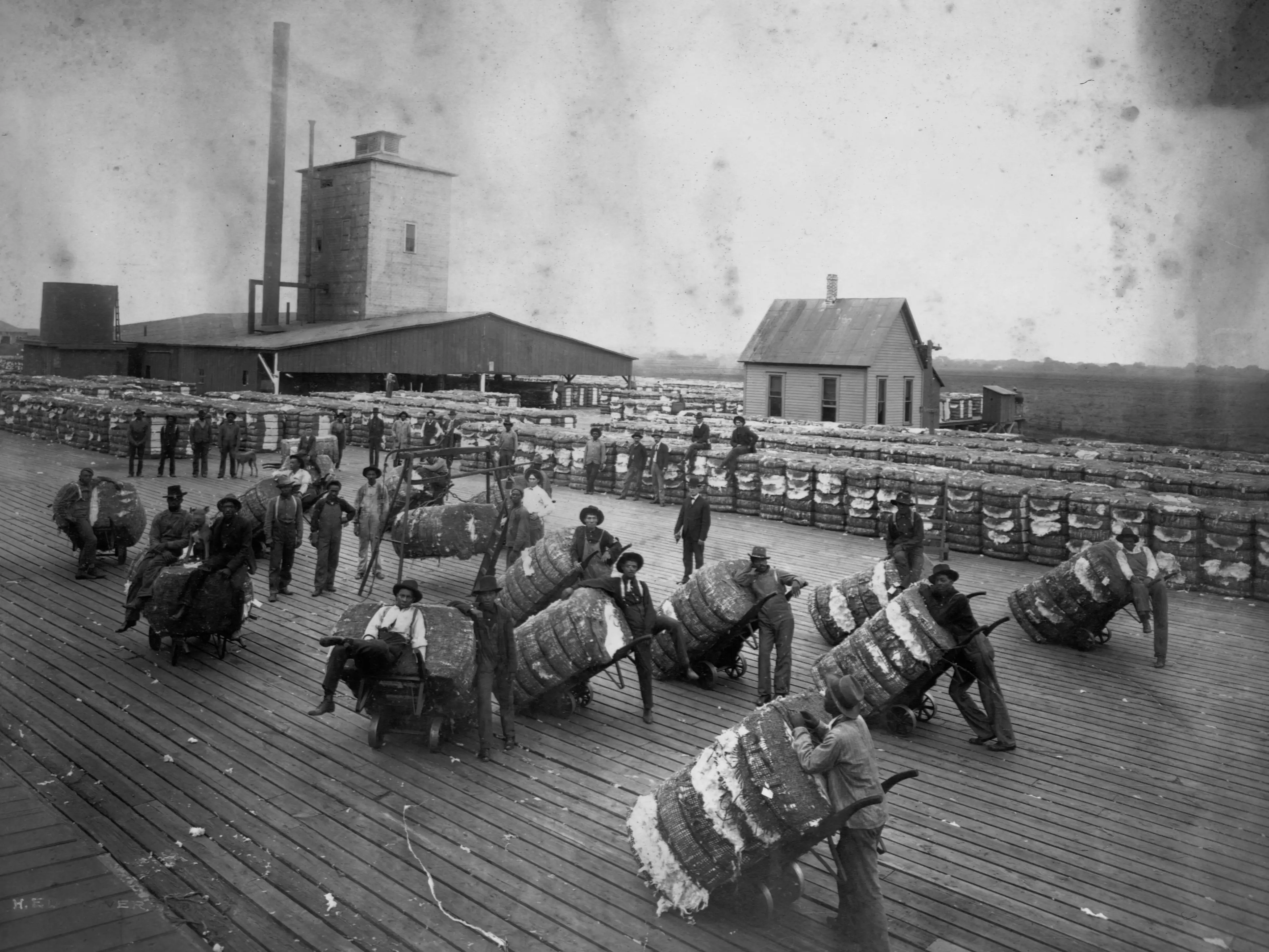 Black men stand with bales of cotton on a loading dock at a cotton compress.