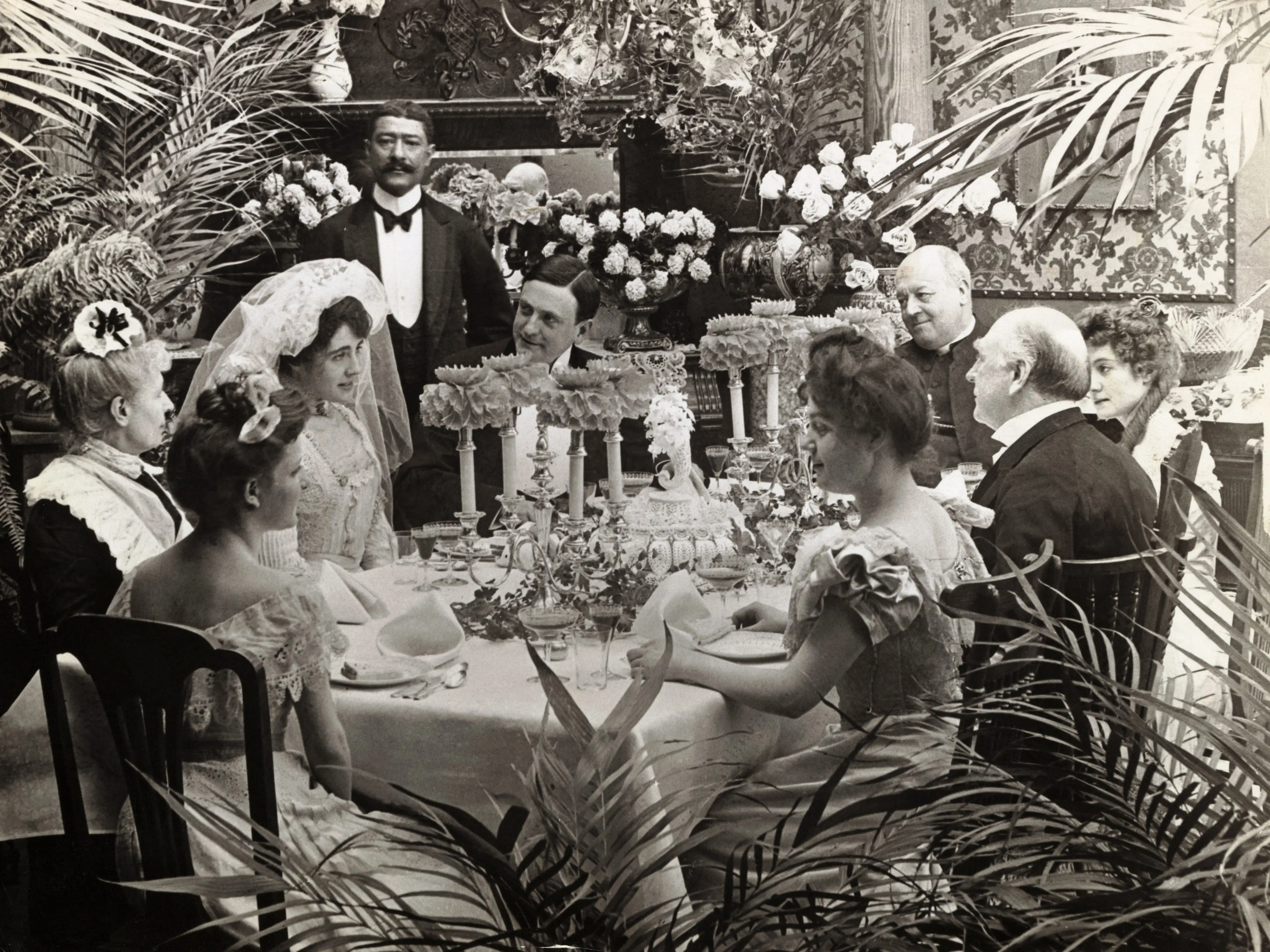 Undated photograph of a wedding reception. Well dressed people formally sitting around a banquet table.