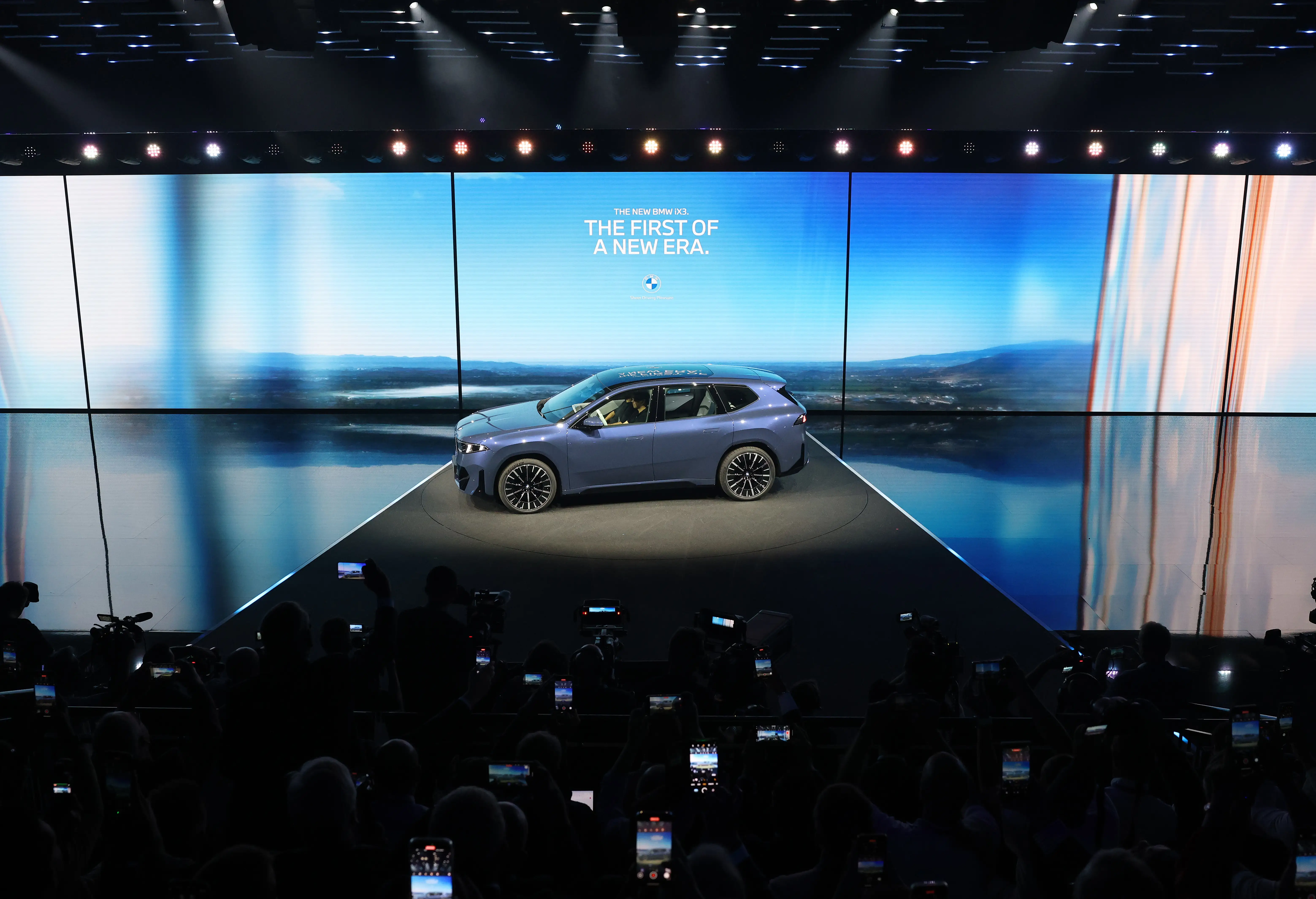 A blue BMW iX3 is parked on a stage in front of a digital sign that says 