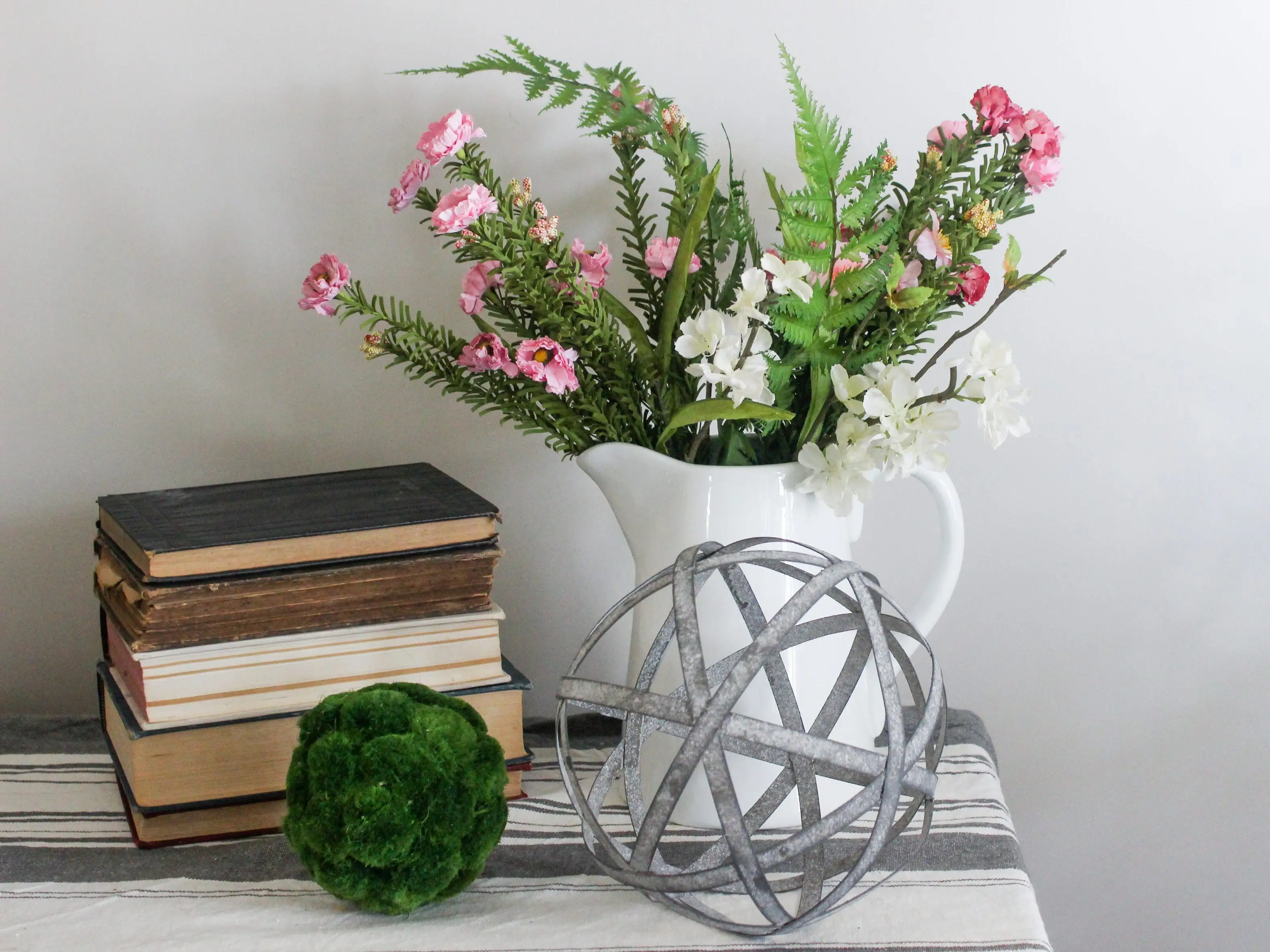 home decor shelf with faux flowers in pitcher, moss ball, stack of books