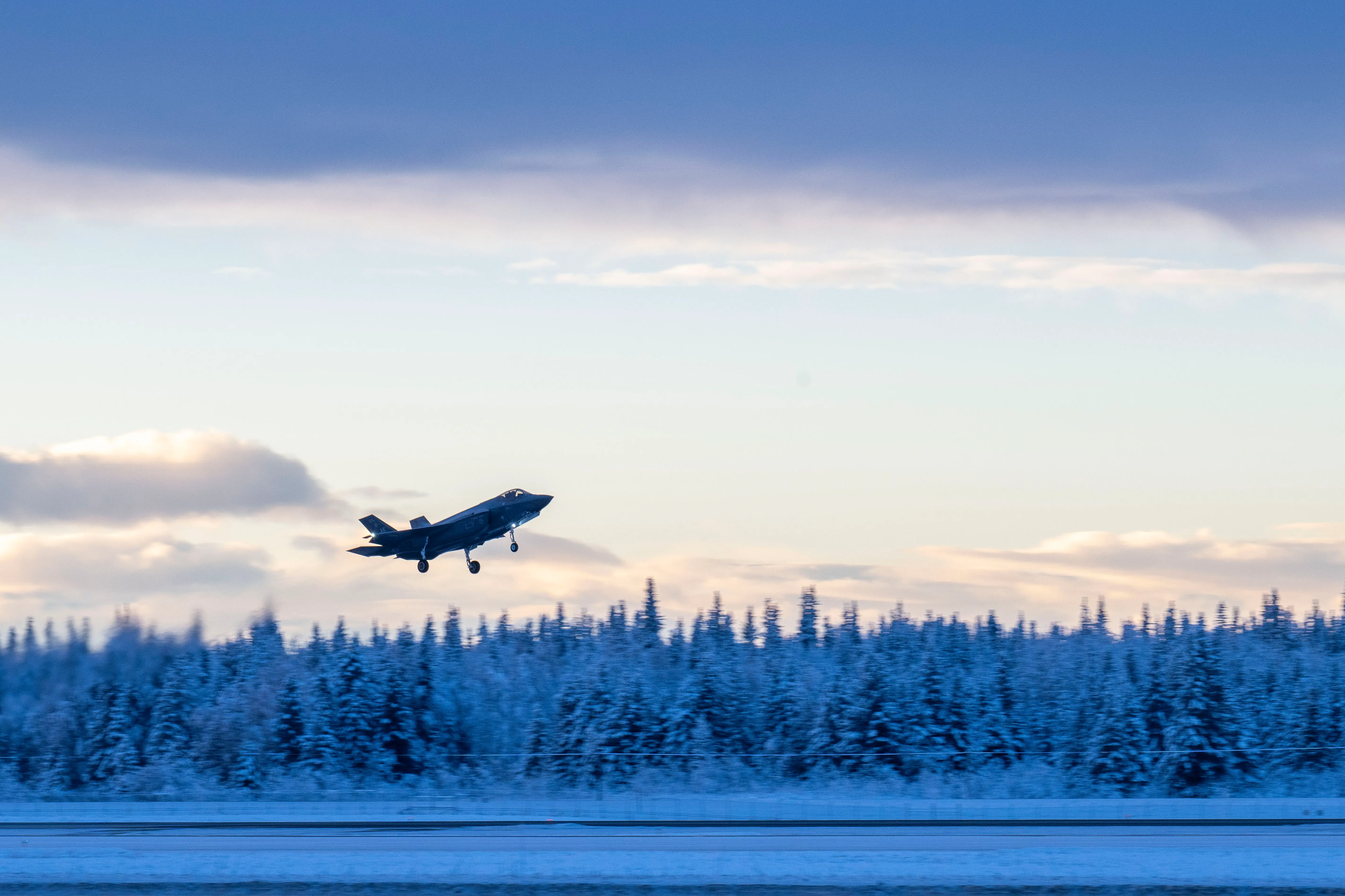 A fighter jet takes off from a snowy landscape with trees and a cloudy sky in the background.