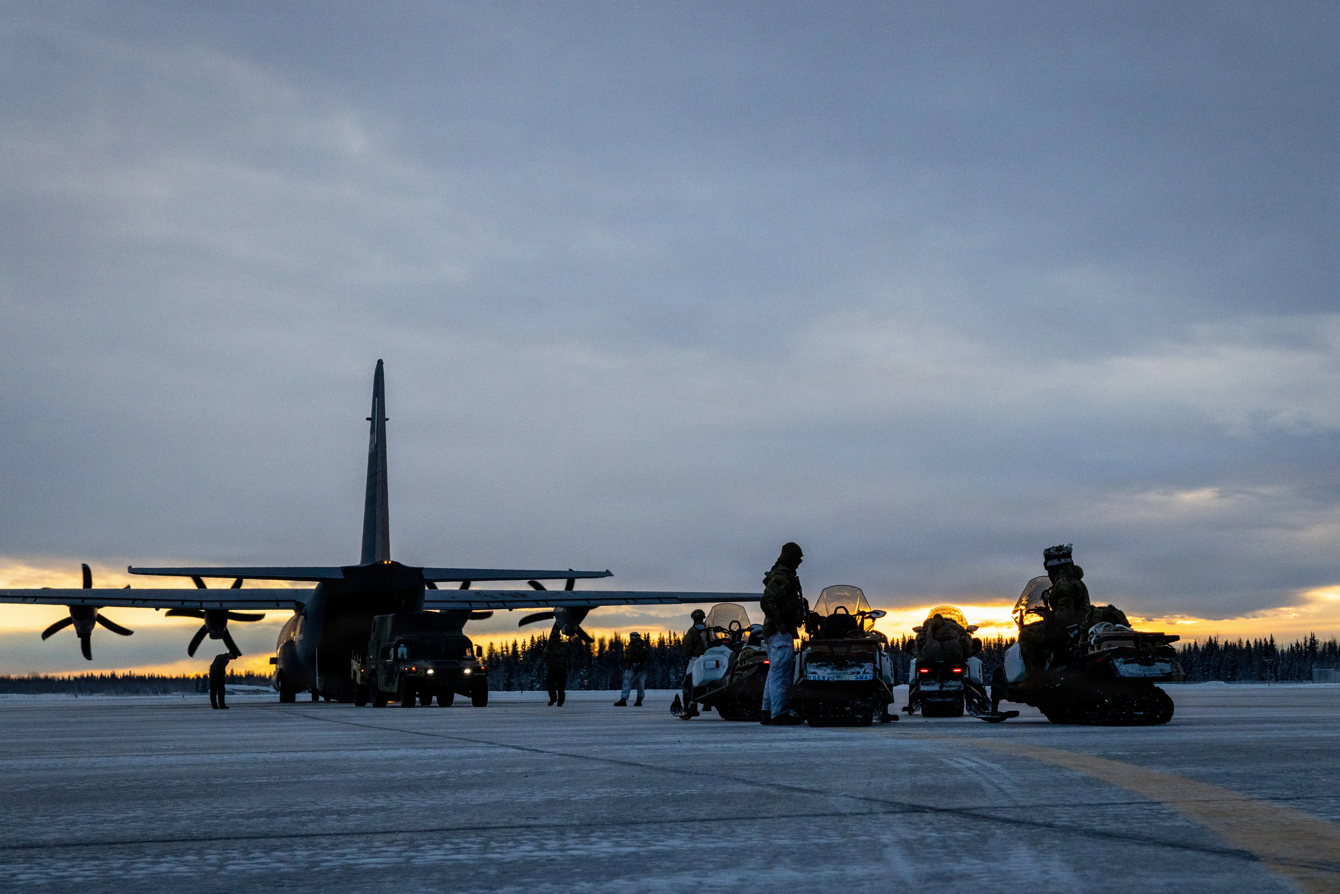 A large military aircraft and several vehicles sit on a tarmac with a cloudy sky in the background.