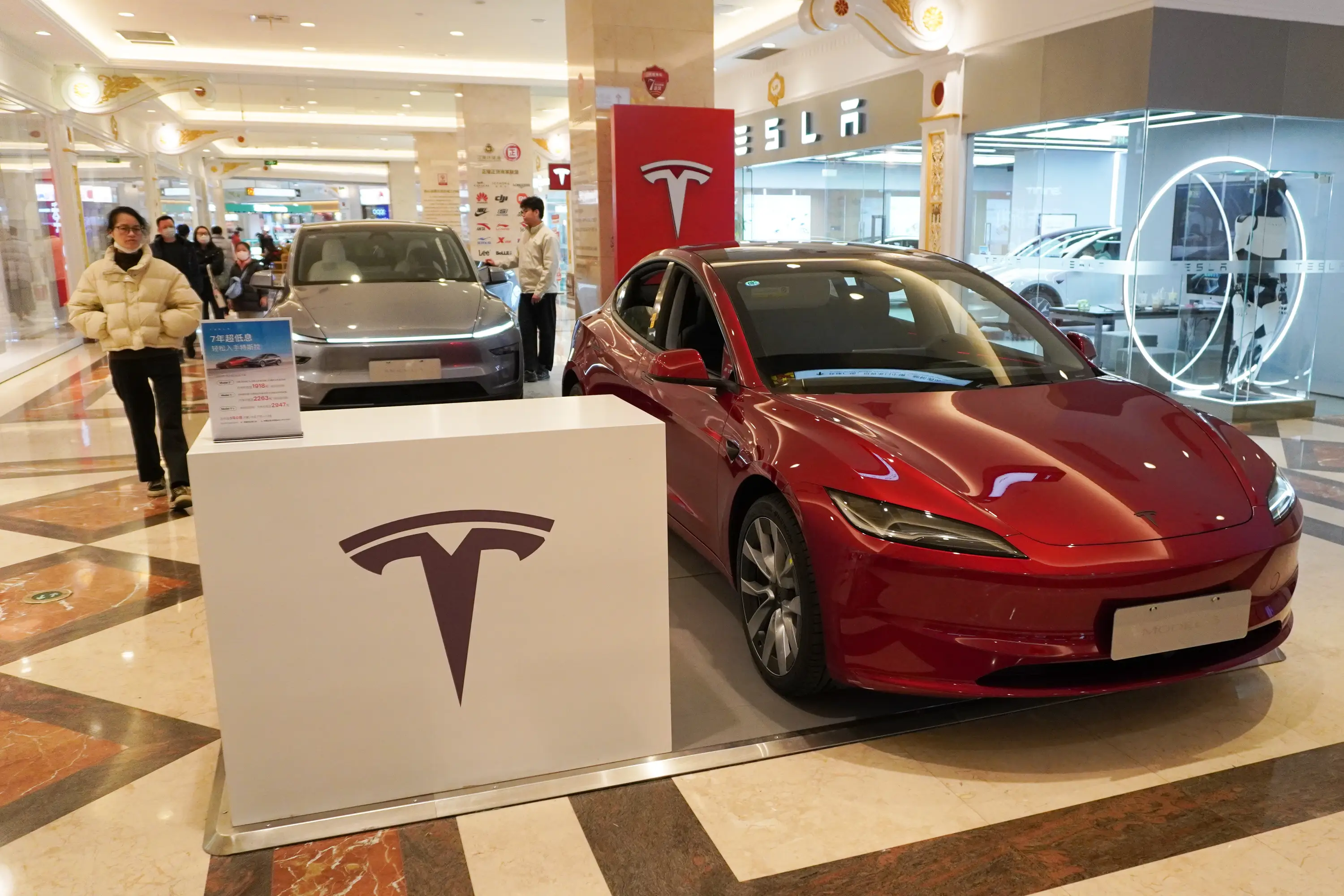 A red Tesla Model 3 and a gray Tesla Model Y seen displayed in the atrium of Global Harbor shopping mall with shoppers passing by.