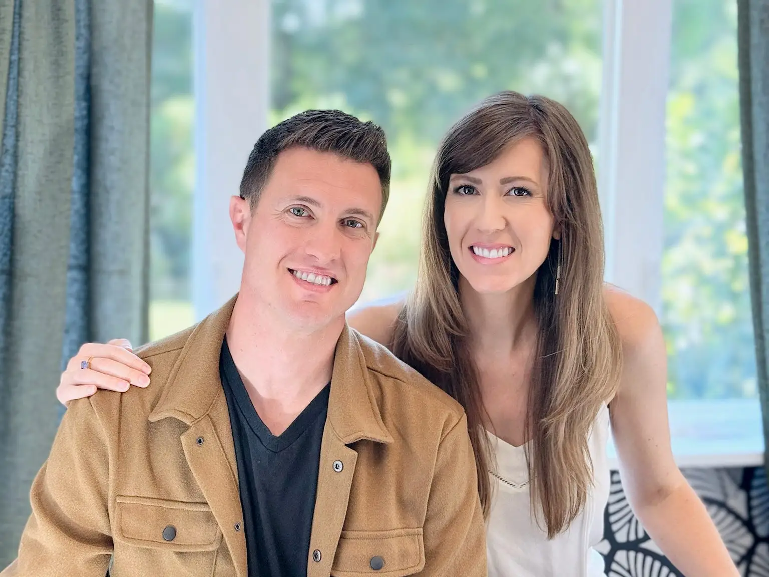 a couple poses for a photo by their window