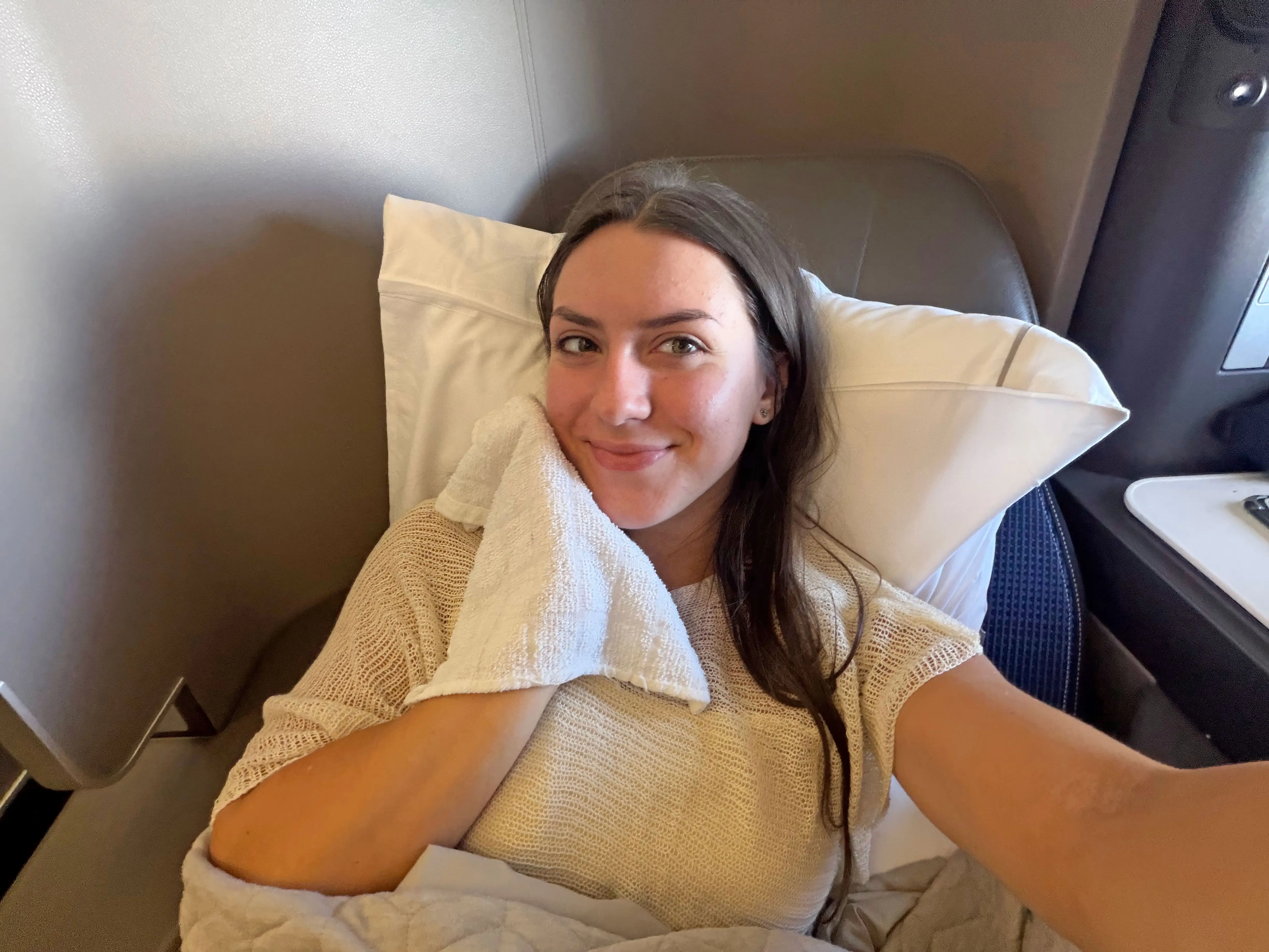 Amanda holds a towel to her face as she lies down in a business-class seat.