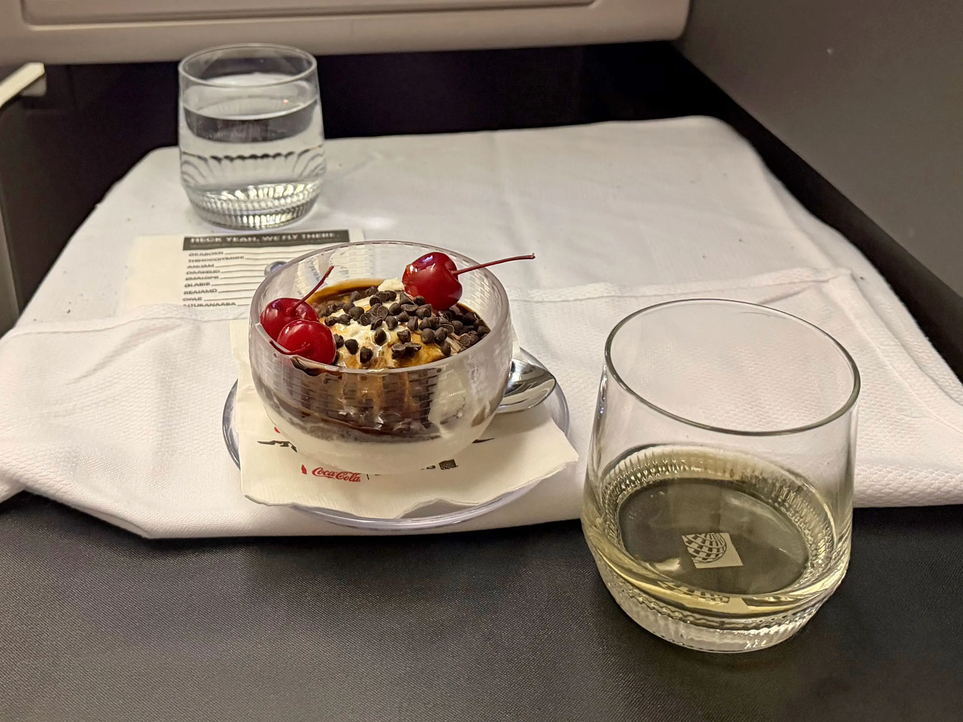 A glass of water, an ice cream sundae, and a glass of white wine on a plane tray table.