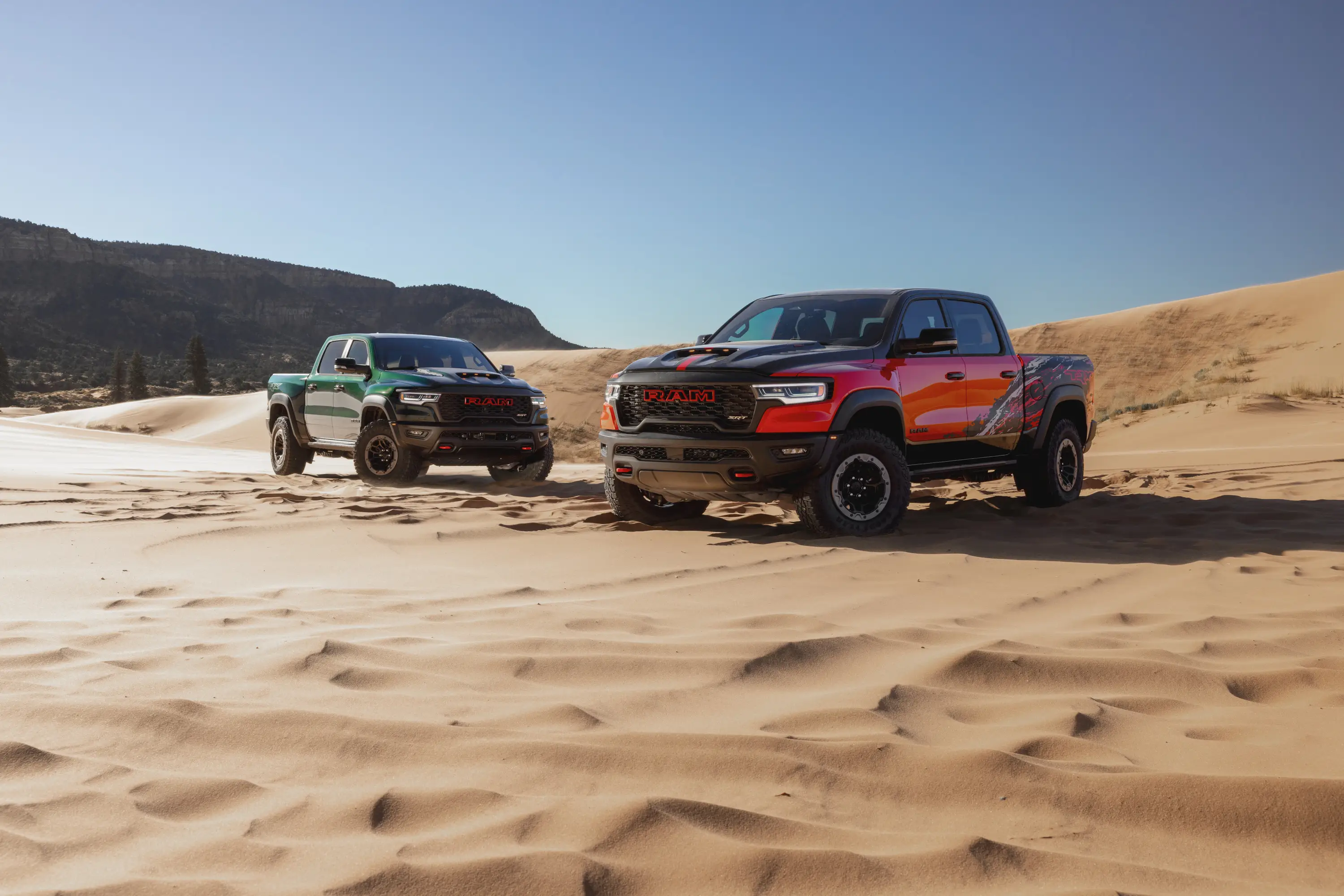 Two Ram pickup trucks are parked on sand dunes. They're both V8-powered TRX models.
