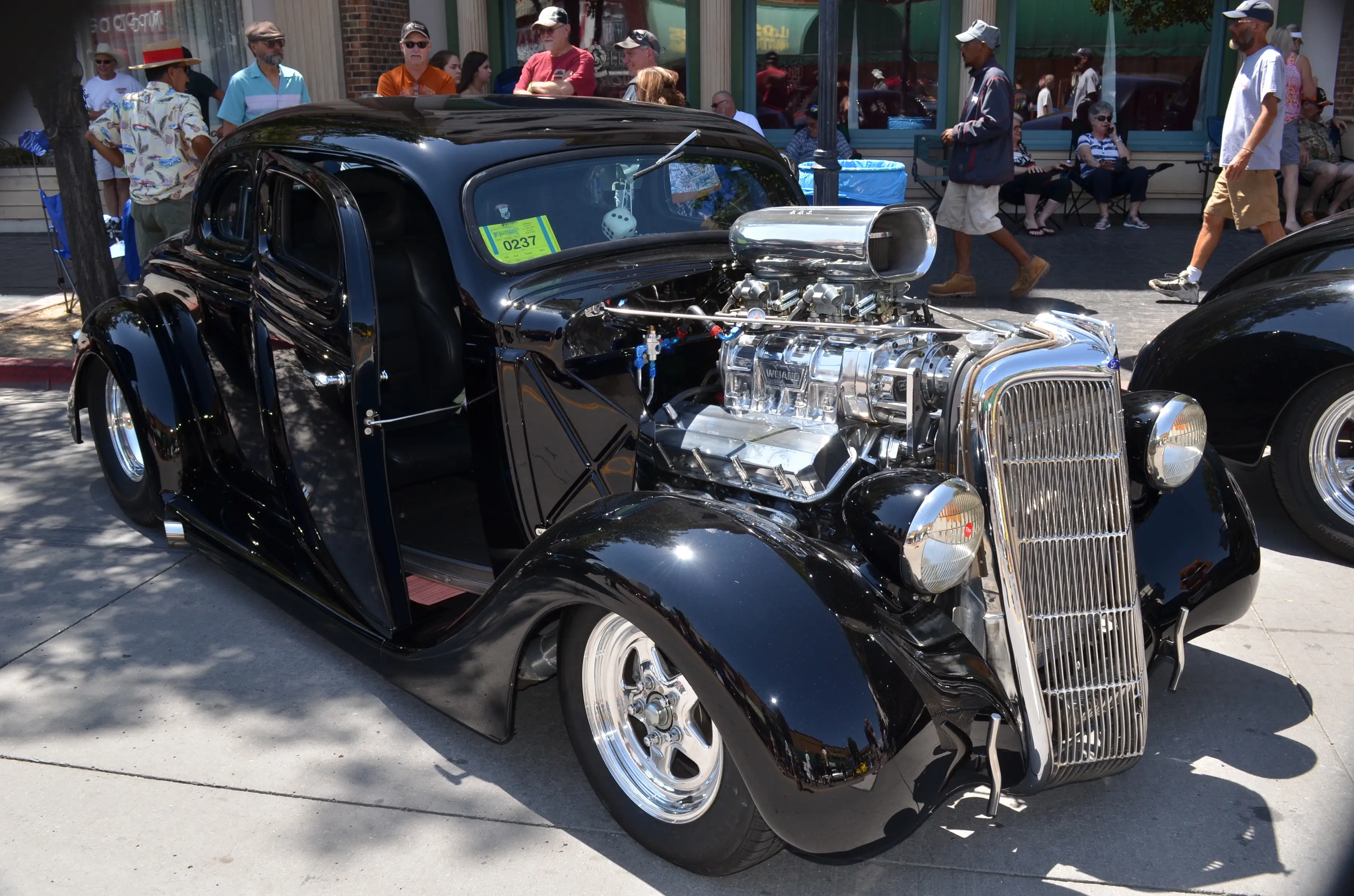 Onlookers mull around a low-riding 1930s Ford Coupe with a massive engine.