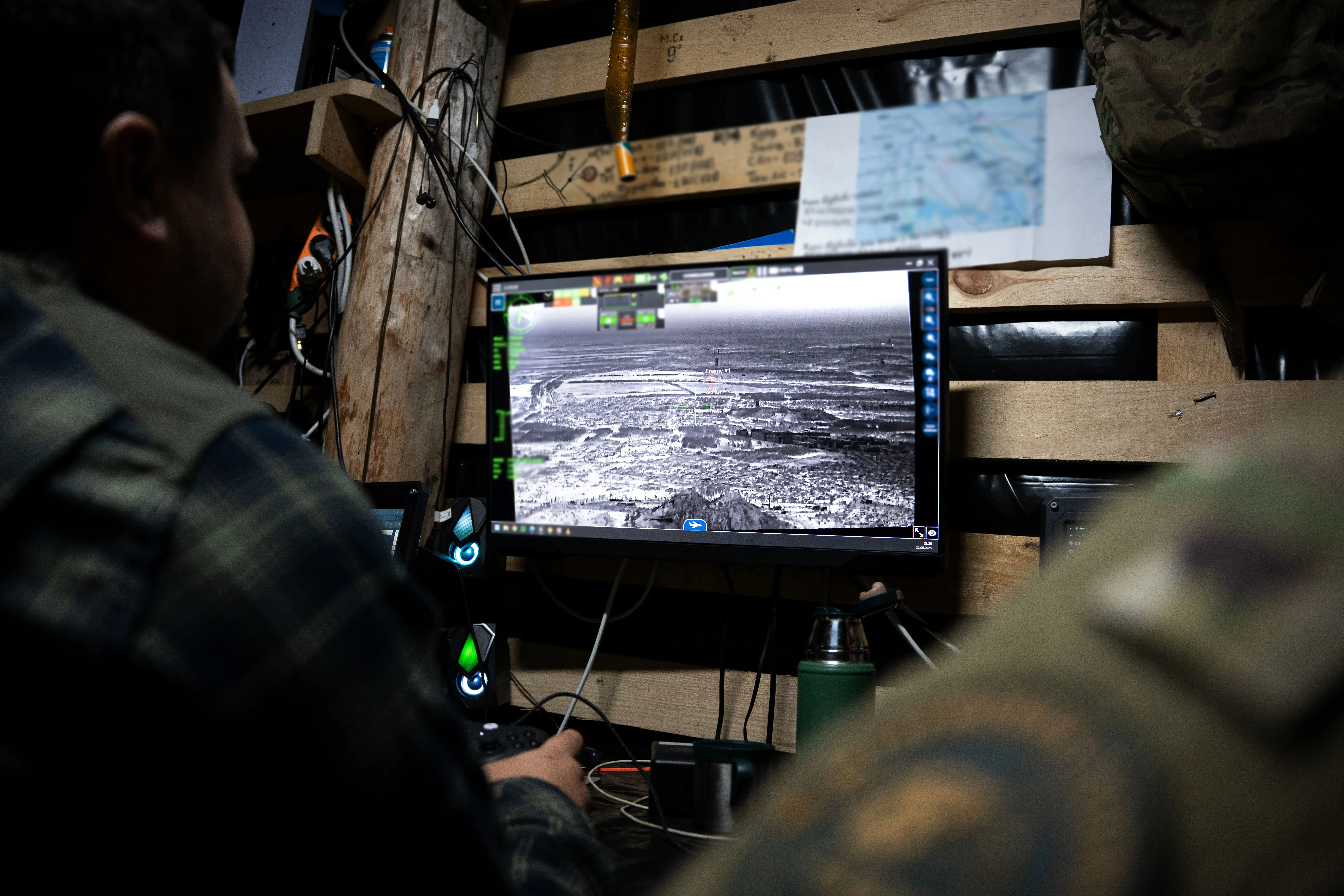 The shoulders of two people sat in front of a monitor displaying an aerial drone field in a dark room with wooden slats behind the monitor