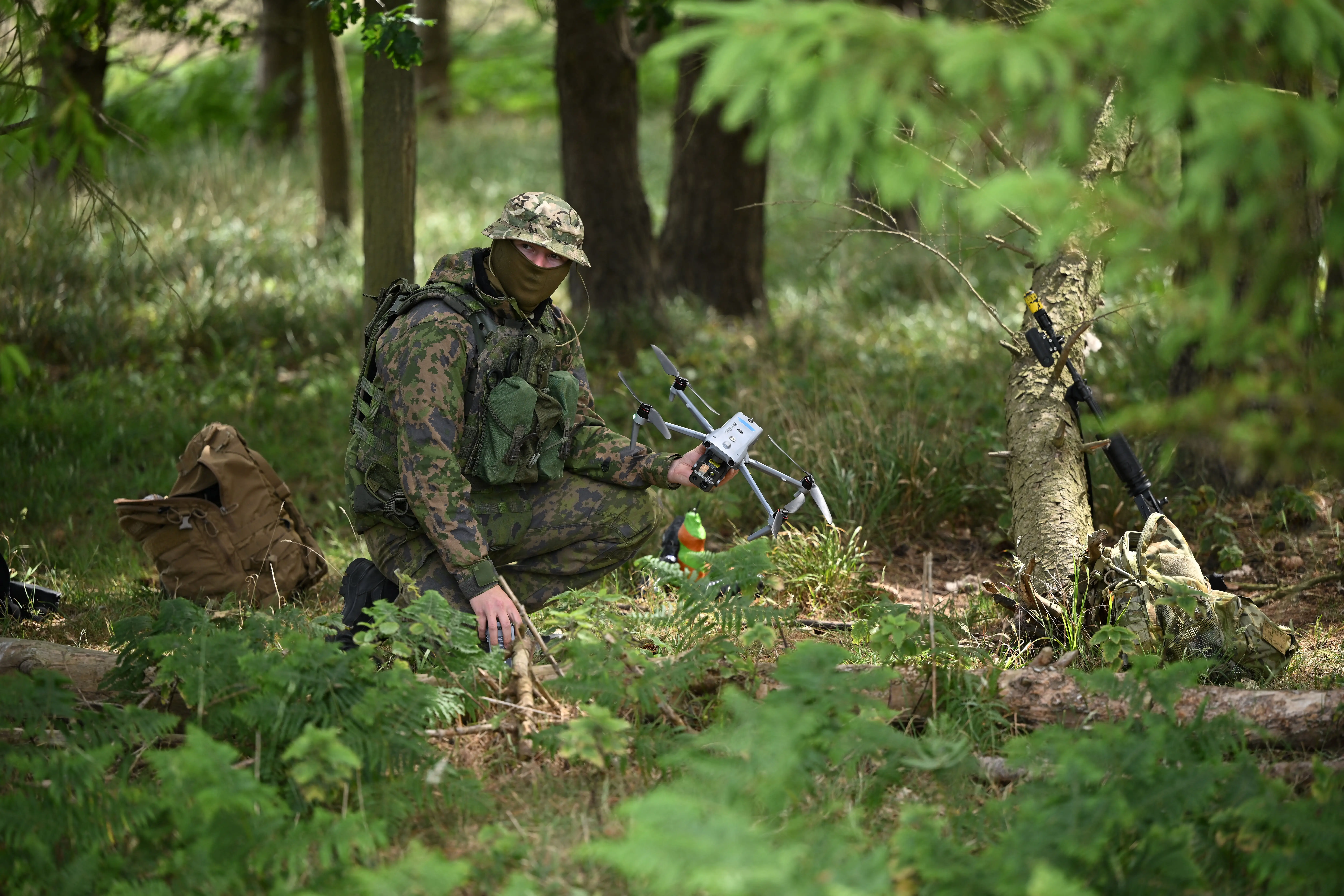 A figure in camouflage squats holding a small grey drone in a wooded area