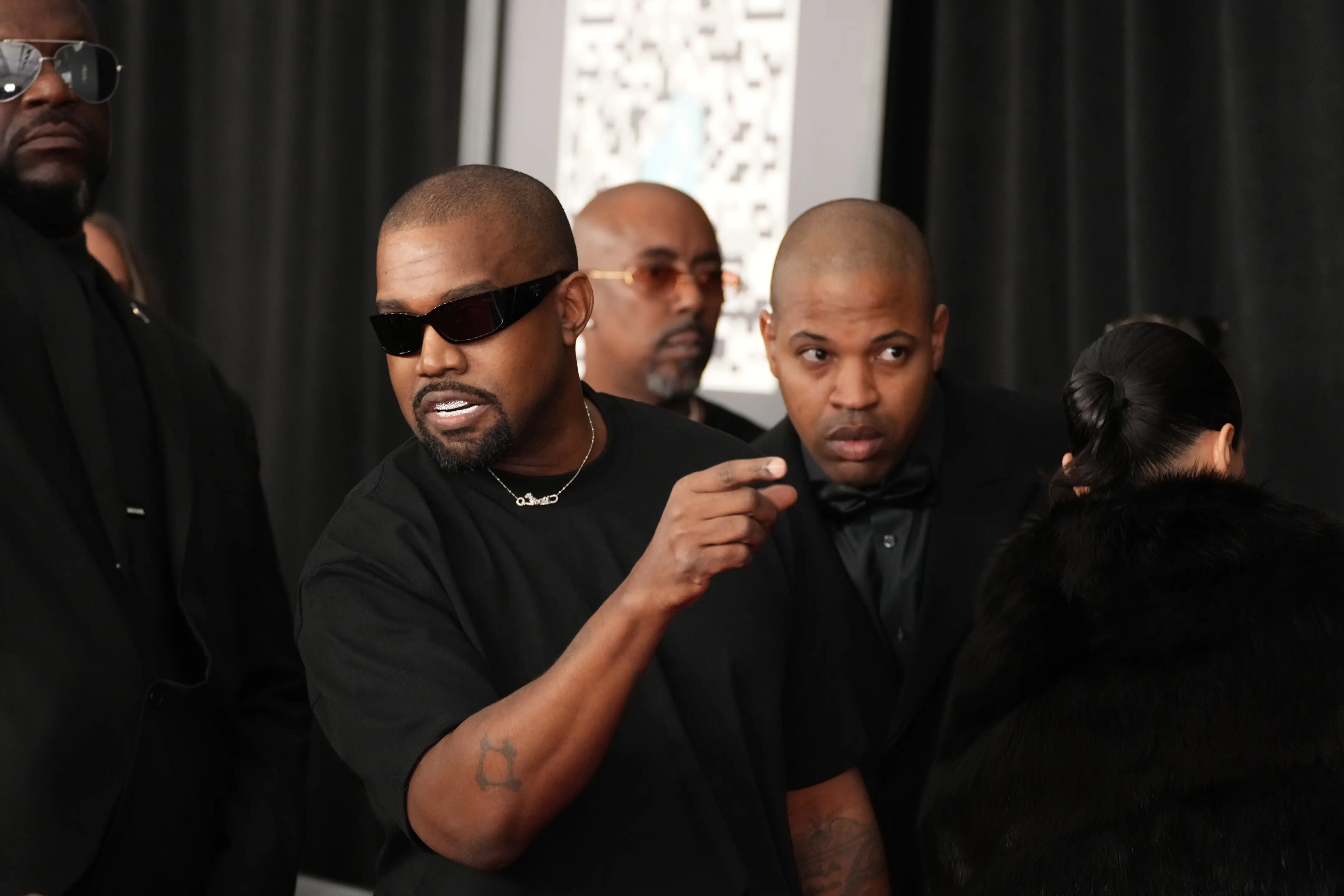 Ye, formerly known as Kanye West, on the red carpet in a black shirt and sunglasses. He's pointing to something to the camera's right.