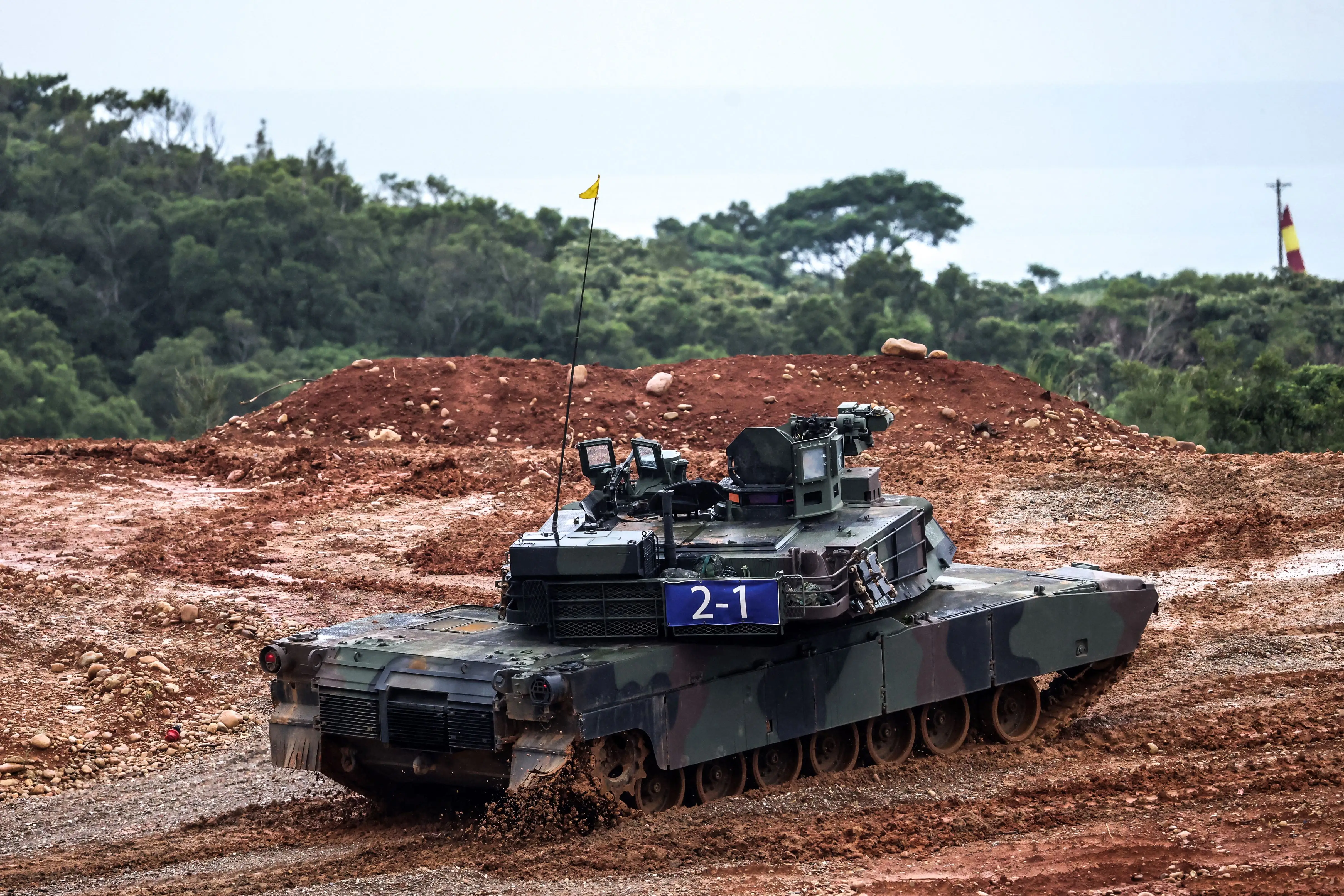 A camouflage-colored M1A2T tank drives in red-colored dirt. There are trees and an overcast sky in the background.