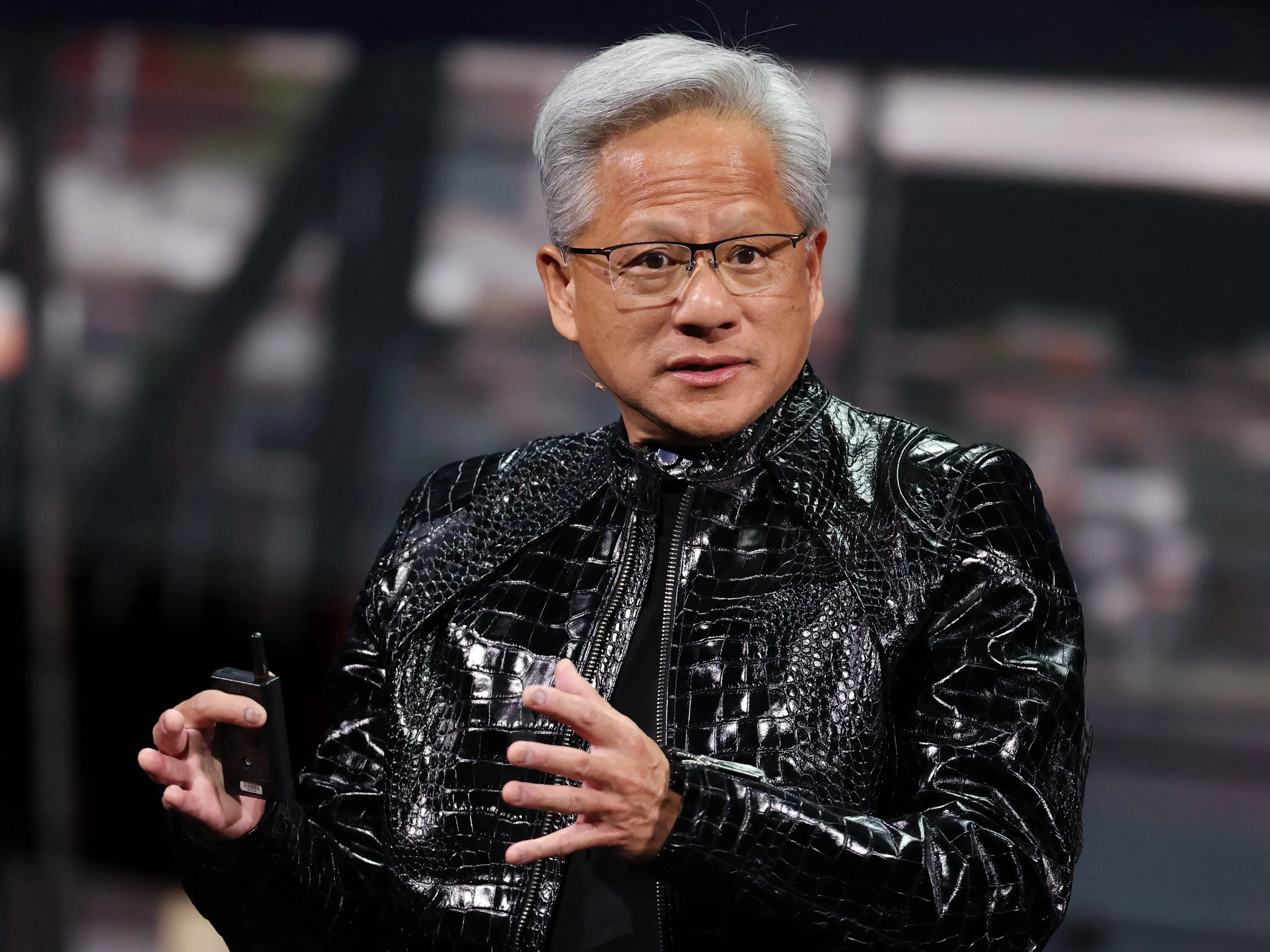 Jensen Huang gesturing with his hands onstage, wearing a black alligator jacket.