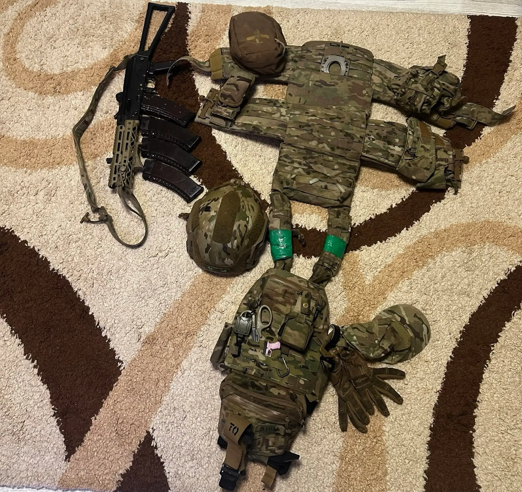 A camouflage-coloured set of body armor and hemlet laid flat, with gear like scissors and gloves visible, and a firearm beside it, all laid on a brown patterned carpet