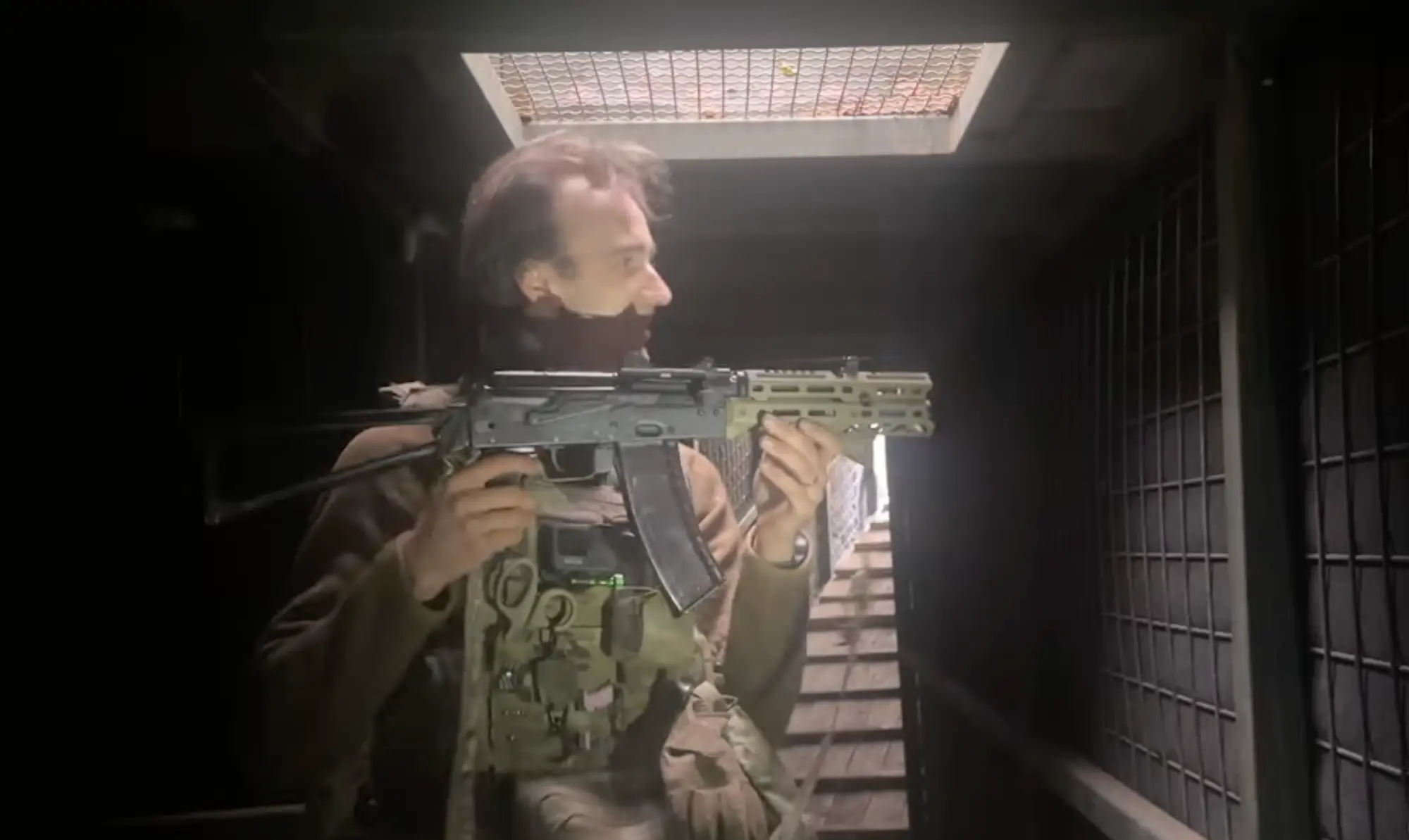 A man in khaki gear stands in an underground position with a mesh skylight and stairs behind him holding a black firearm with a green handguard