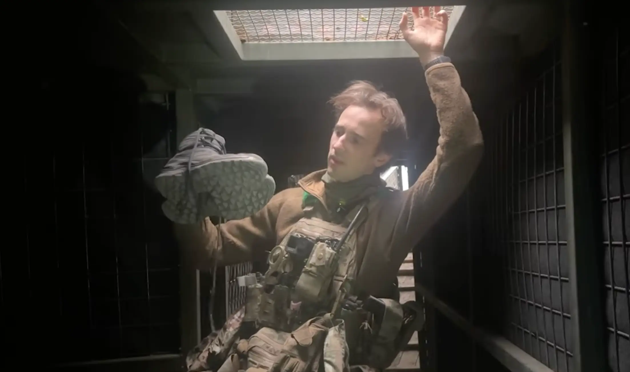 A man in khaki gear stands in an underground position with a mesh skylight and stairs behind him holding a pair of grey boots up with one hand