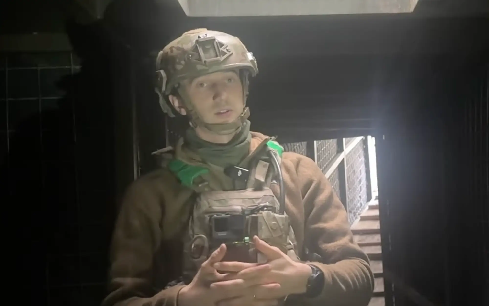 A man in khaki gear stands in an underground position with stairs behind him stands wearing a helmet